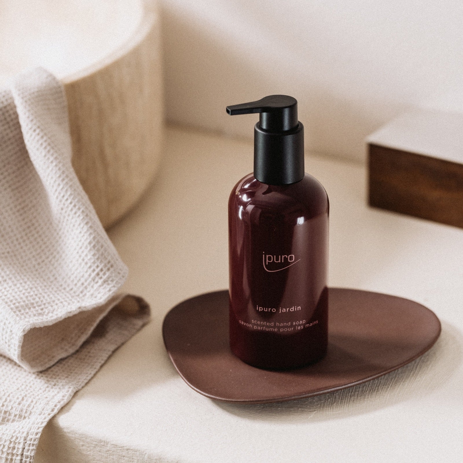 Eine bernsteinfarbene Pumpflasche mit CLASSIC ipuro jardin Handseife von ipuro ruht auf einem braunen Tablett neben einem cremefarbenen Handtuch und einem runden beigen Waschbecken und verleiht der minimalen, sanft beleuchteten Szene einen Hauch von exotischen Blüten.