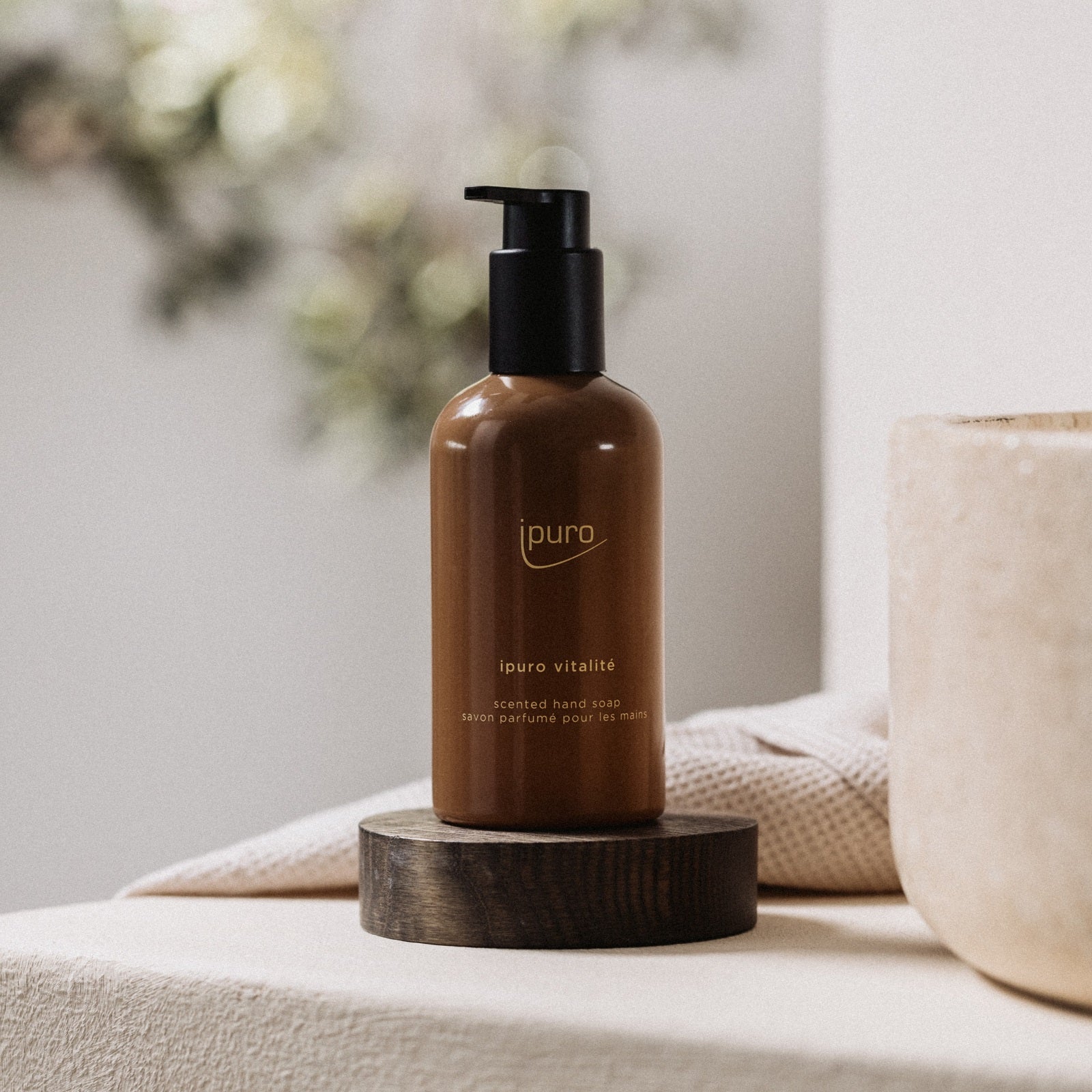 Eine braune Pumpflasche der ipuro CLASSIC vitalité Handseife steht auf einem Holzuntersetzer neben einem beigen Handtuch. Das neutrale Dekor erinnert an eine Spa-Atmosphäre und lässt den erfrischenden Rosmarin-Ingwer-Duft der Seife erahnen.
