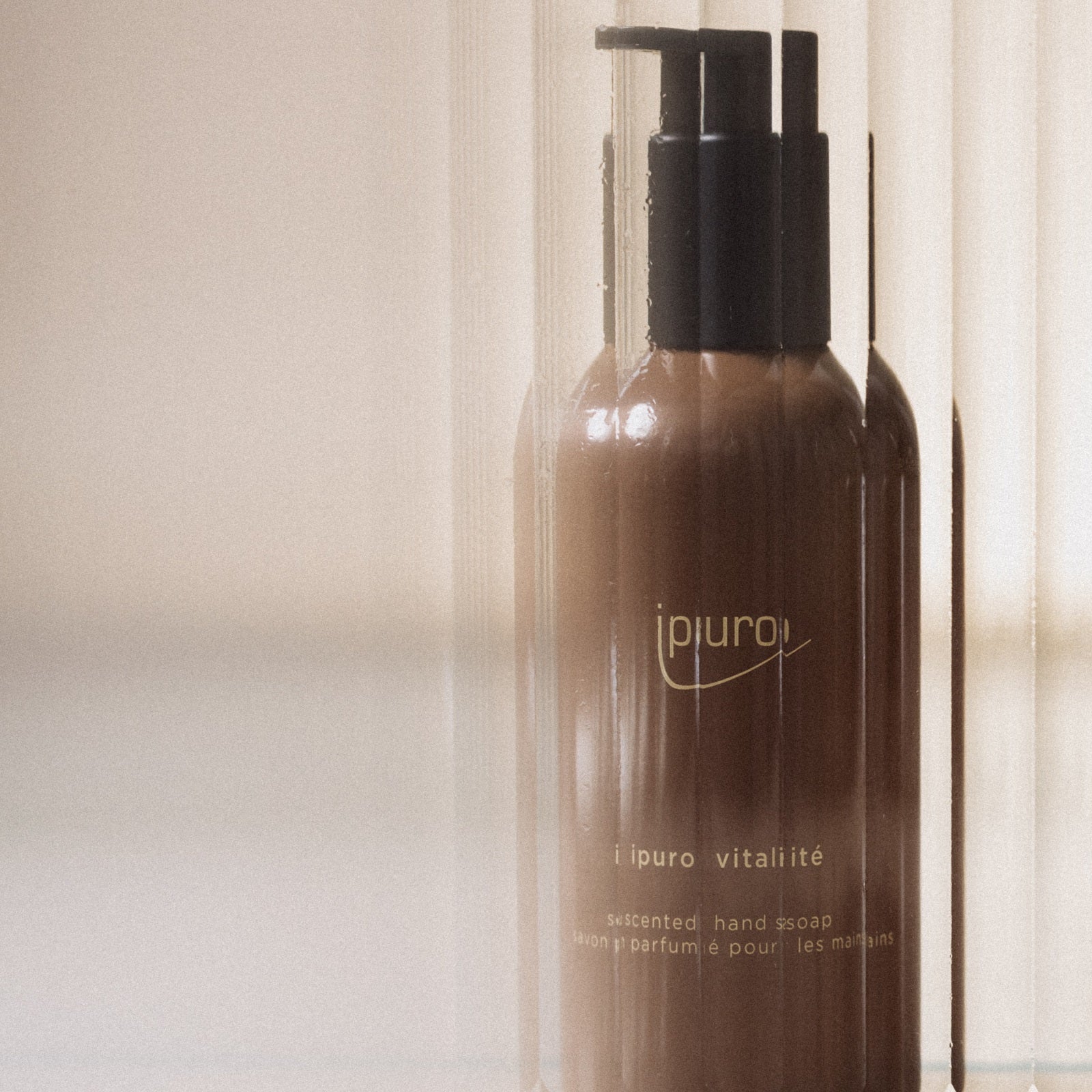 Eine braune Pumpflasche mit CLASSIC ipuro vitalité Handseife von ipuro steht vor einem weichen, verschwommenen Hintergrund mit Doppelbelichtungseffekt, der an Wellness und den erfrischenden Duft von Rosmarin und Ingwer erinnert.