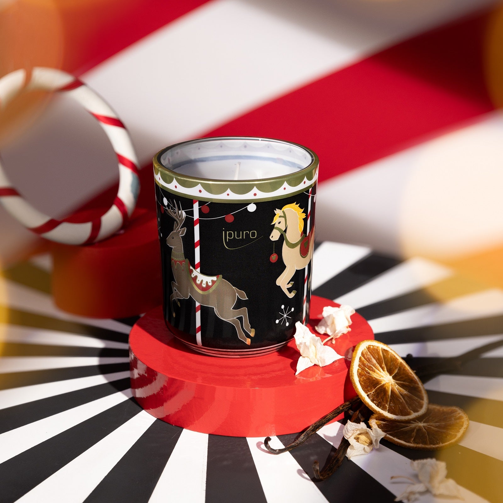 Die LIMITED EDITION ipuro magical market Kerze 270gr von ipuro ist eine dekorative Kerze in einem festlichen Halter mit Karussellpferd- und Rentiermotiven, auf einem roten Ständer, akzentuiert durch getrocknete Orangenscheiben und weiße Blütenblätter vor einem gestreiften Hintergrund.