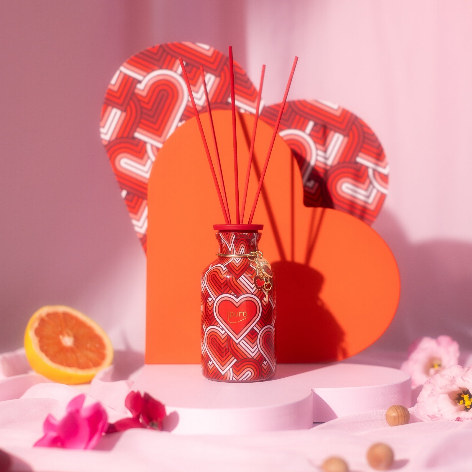 Der LIMITED EDITION ipuro with love Raumduft Diffusor von ipuro mit rotem und weißem Herzmuster ist zwischen Blumen, rosa Dekor und einer Orangenscheibe auf einer zartrosa Fläche vor großen herzförmigen Kulissen abgebildet.