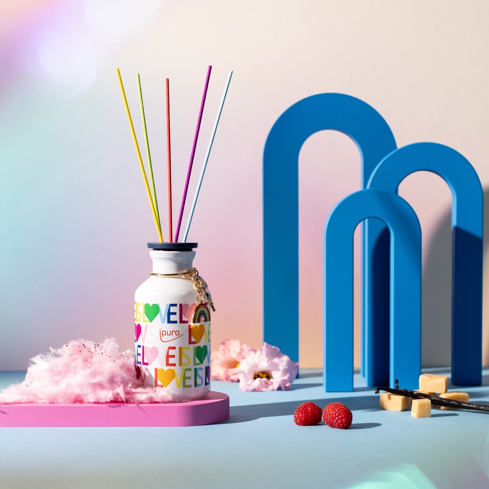 Der LIMITED EDITION ipuro love is love Raumduft von ipuro, ein farbenfroher Schilfdiffusor mit Regenbogenstäbchen und herzverzierter Flasche, sitzt auf pastellfarbenen Bögen, rosa Zuckerwatte und Himbeeren und versprüht einen Hauch von Erdbeersüße.