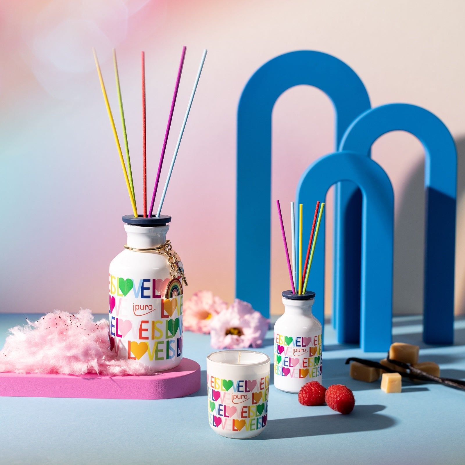 Die LIMITED EDITION ipuro love is love Raumduft von ipuro, gepaart mit einer Kerze, Himbeeren, Vanille-Karamellwürfeln und rosa Zuckerwatte, wird auf pastellfarbenen Oberflächen mit kräftigen blauen Bögen für eine verspielte Atmosphäre präsentiert.