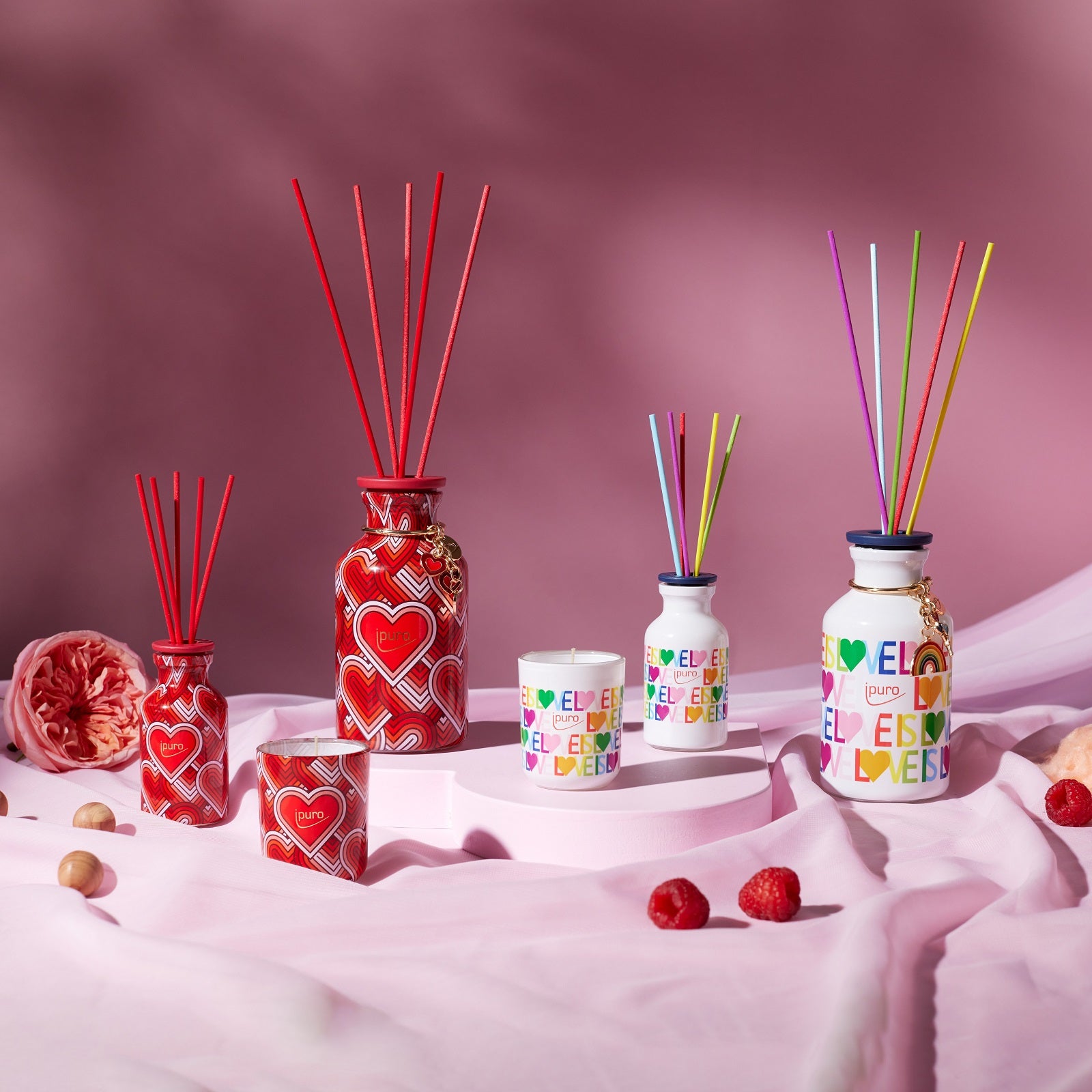 Eine Kollektion von farbenfrohen ipuro LIMITED EDITION "love is love" Raumduft Diffusoren und Kerzen in Herzform, beduftet mit Zuckerwatte & Erdbeere, arrangiert auf rosa Stoff mit Himbeeren und einer Rose, von ipuro.