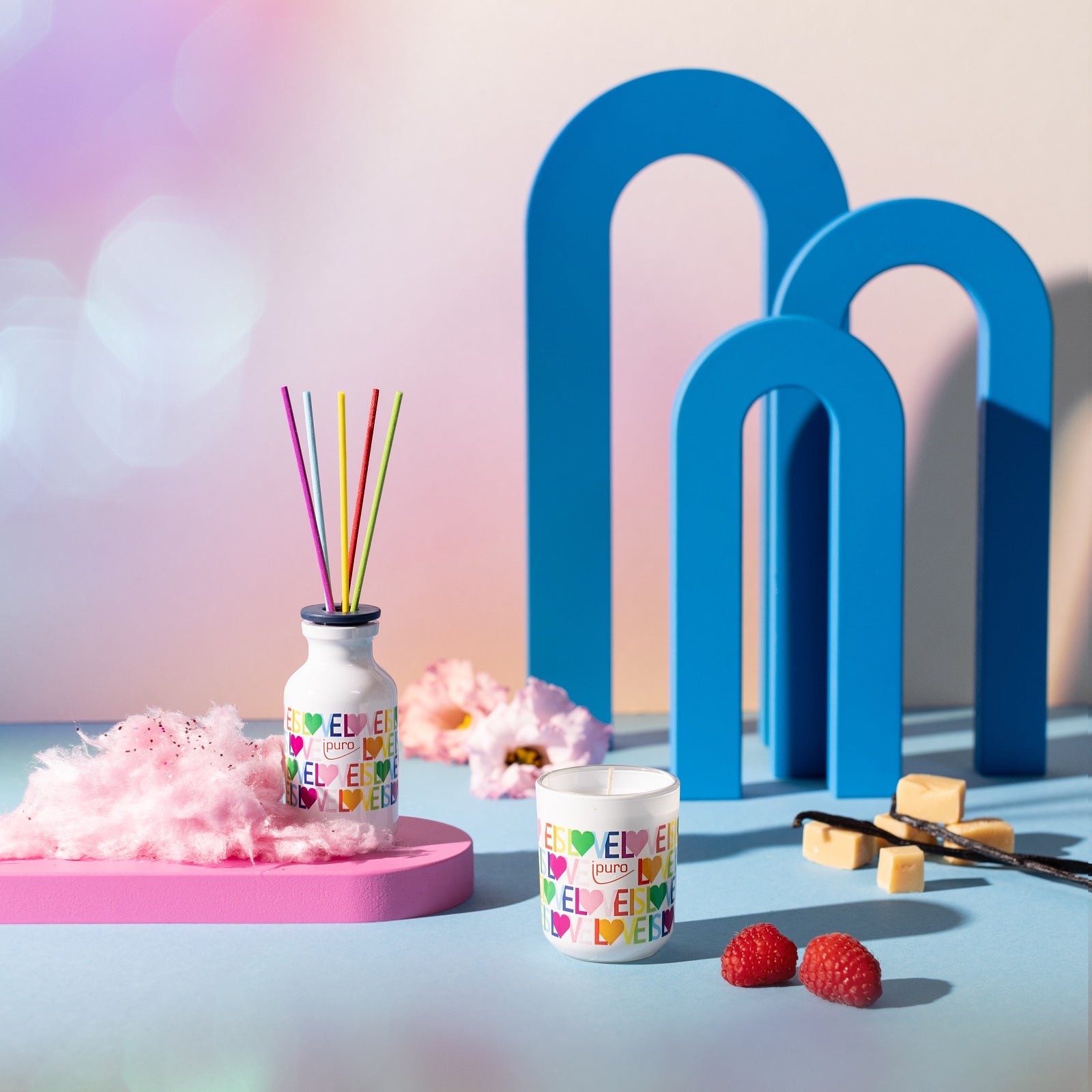 Das ipuro love is love Geschenkset in der LIMITED EDITION von ipuro zeigt eine lebhafte Pastell-Szene mit einem Diffusor und einer Duftkerze inmitten von Zuckerwatte, Himbeeren, Karamell und verspielten blauen Bögen für einen süßen Vanille-Gourmand-Duft.