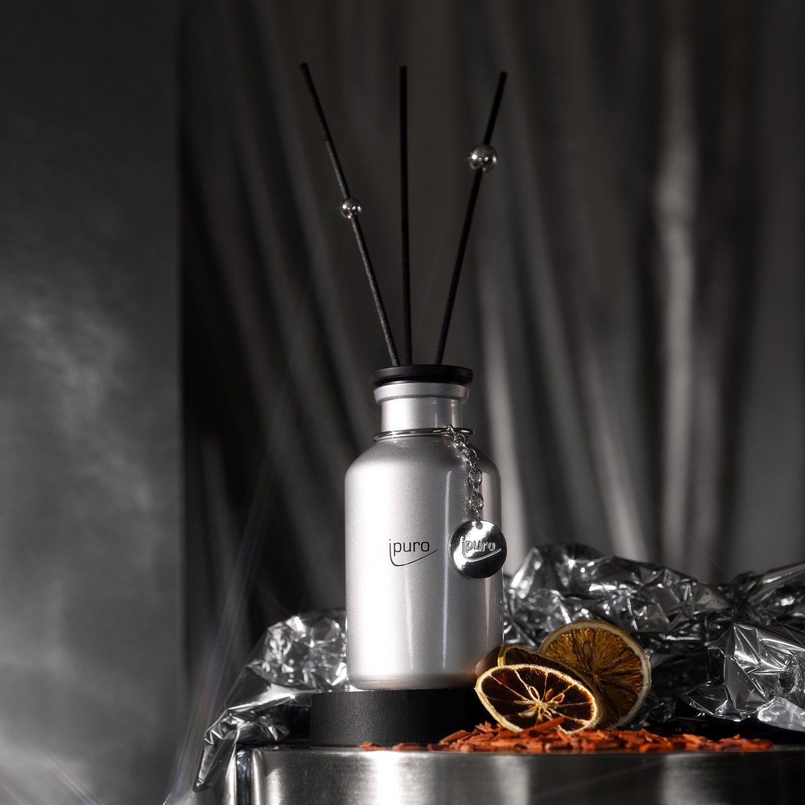 Der LIMITED EDITION ipuro cold glamour Raumduft von ipuro mit schwarzem Schilf liegt zwischen getrockneten Orangenscheiben, Blütenblättern und Metallfolie mit warmen Kaschmir- und Zedernholznoten vor einem verschwommenen dunklen Hintergrund.