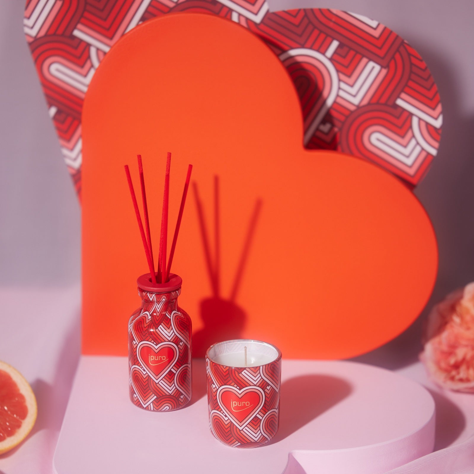Das LIMITED EDITION ipuro with love Geschenk-Set von ipuro besteht aus einer roten, herzförmigen Schachtel, einem passenden Diffusor und einer Duftkerze mit Herzmotiven, die mit einer Grapefruitscheibe und einer Blume für einen blumigen Duft auf Rosa präsentiert wird.