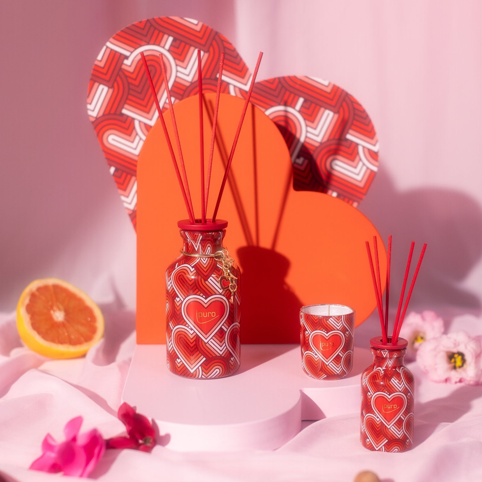 Das LIMITED EDITION ipuro with love Geschenk-Set von ipuro besteht aus farbenfrohen Schilfrohrdiffusoren und einer Kerze mit rotem Herzmuster, die auf herzförmigen Ständern zwischen Blumen und einer Grapefruitscheibe für ein romantisches, blumiges Ambiente sorgen.