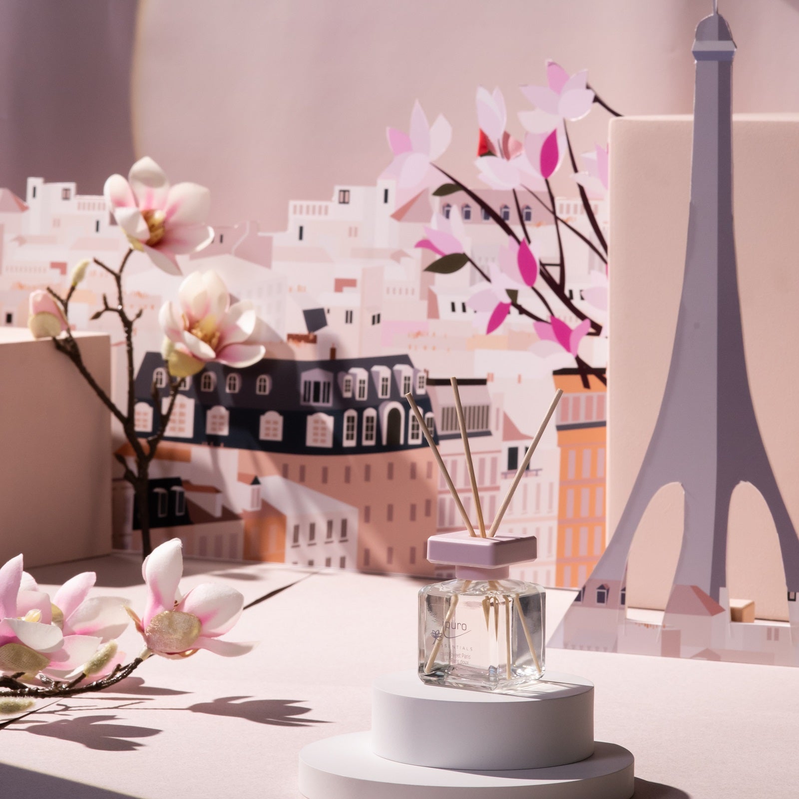 Eine ESSENTIALS ipuro sweet paris Raumduft-Flasche von ipuro steht auf einem runden weißen Ständer, umgeben von rosa Magnolienblüten mit einer pastellfarbenen Pariser Stadtlandschaft und dem Eiffelturm im Hintergrund.