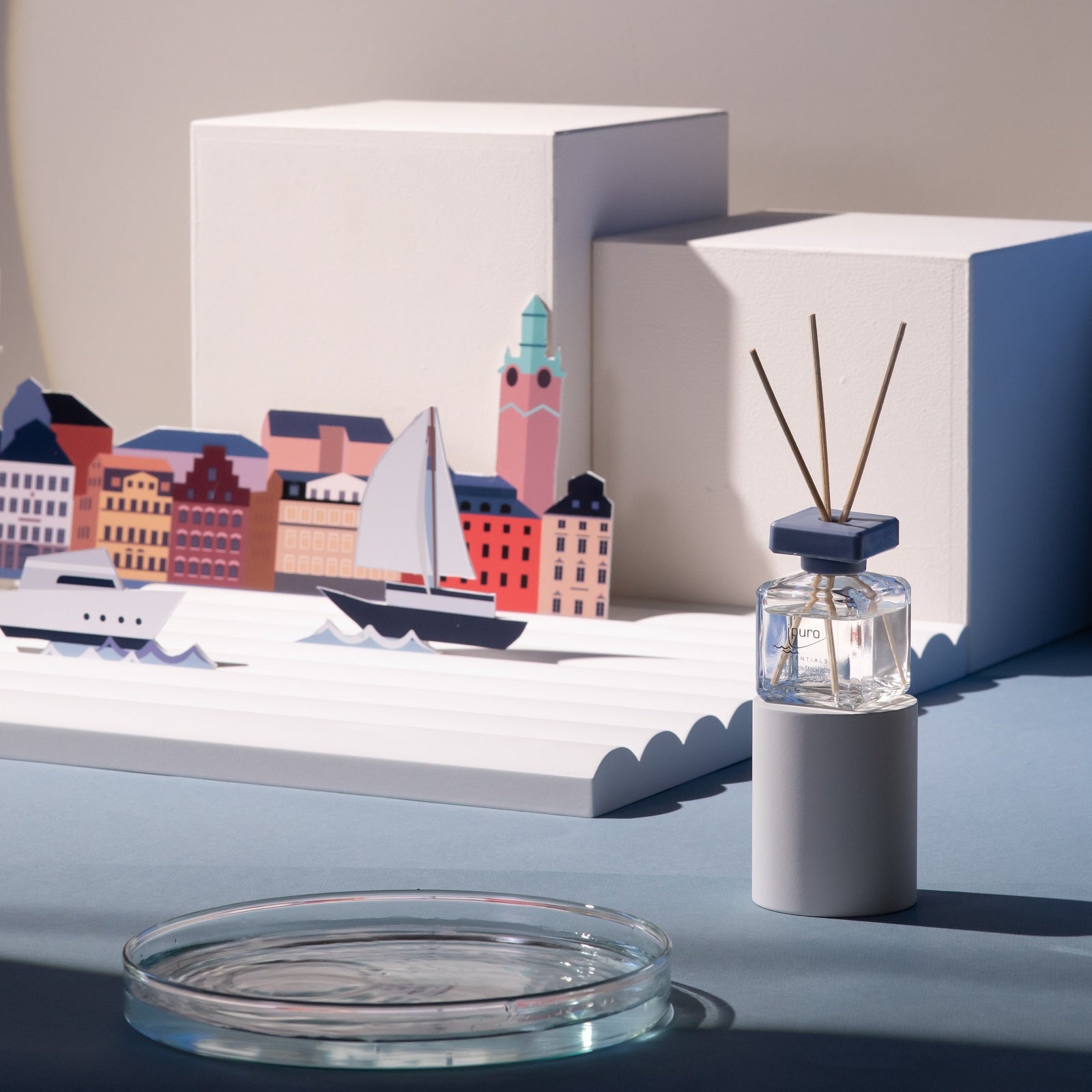 Der ESSENTIALS ipuro fresh stockholm Raumduft von ipuro steht auf einem weißen Ständer vor einer vom Stockholmer Hafen inspirierten Kulisse mit bunten Ausschnitten von Gebäuden und Booten am Meer, die auf maritime Düfte und frische Meeresakkorde hinweisen.