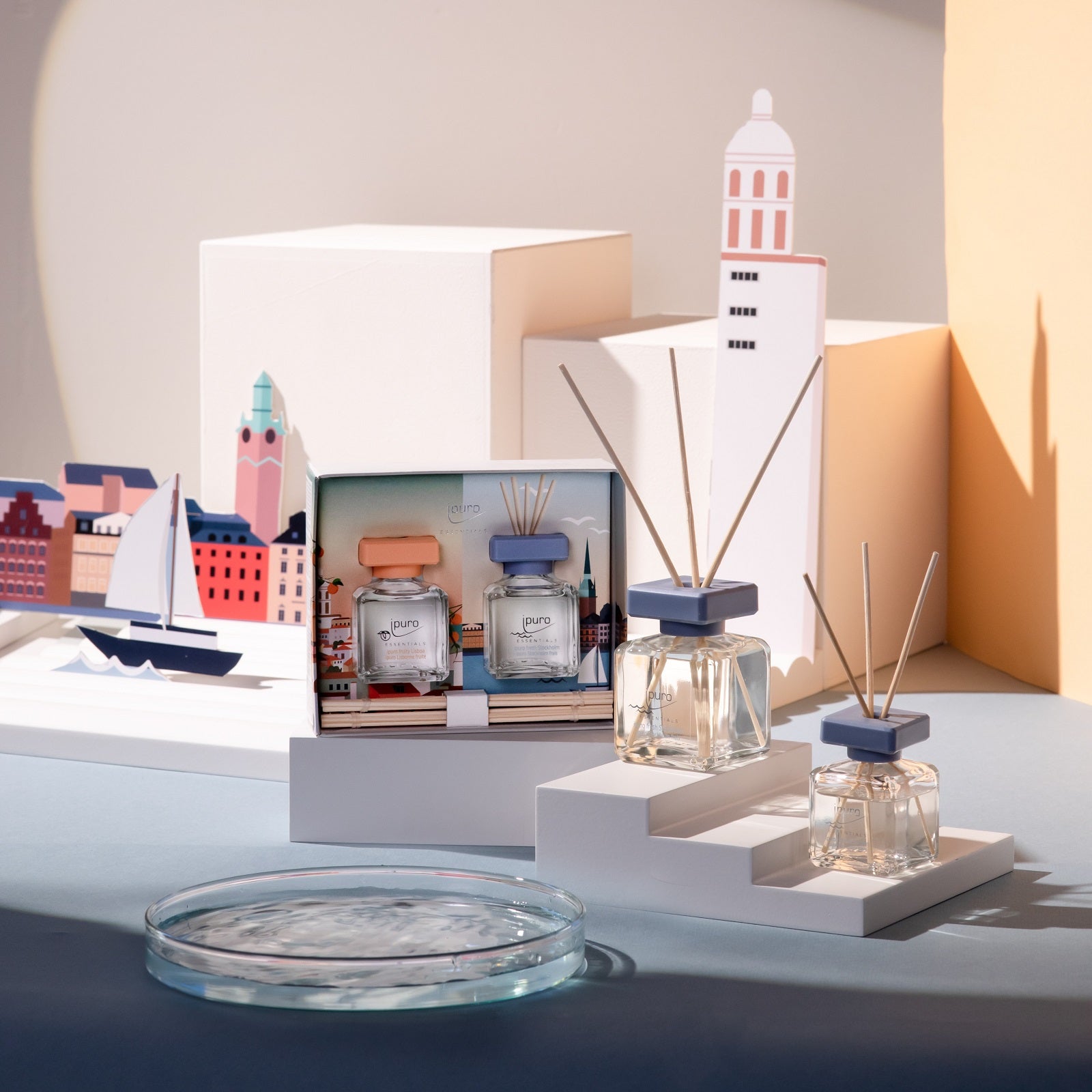 Der ESSENTIALS ipuro fresh stockholm Raumduft von ipuro und das Geschenkset werden auf geometrischen, weißen Plattformen mit Aussparungen für Stadtansichten und Segelboote präsentiert und spiegeln einen modernen, eleganten Stil wider, der vom Stockholmer Hafen und frischen maritimen Aromen inspiriert ist.