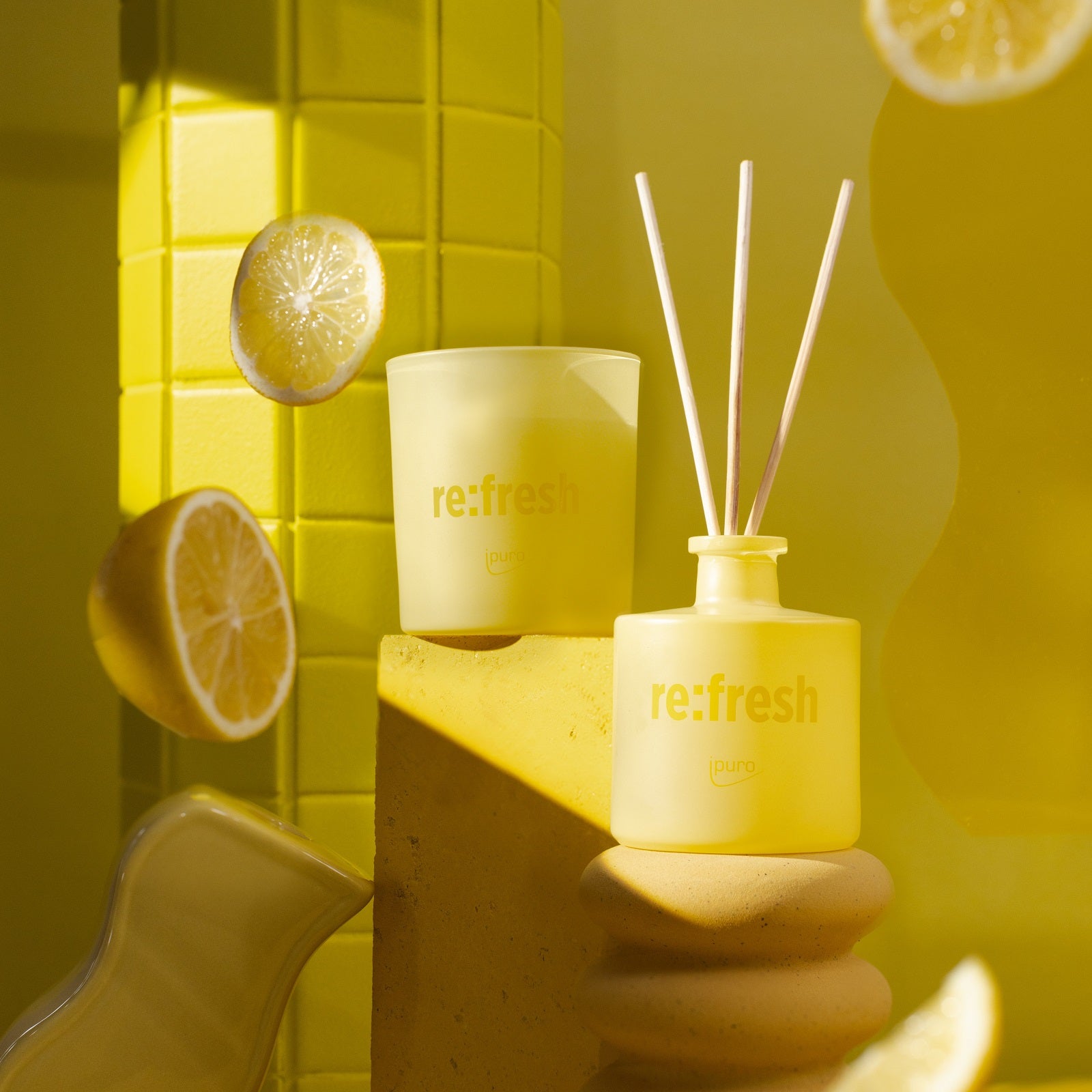 Ein von Gelb inspiriertes Flatlay mit dem ipuro WE ARE: re:fresh Raumduft von ipuro, einer Kerze, schwebenden Zitronenscheiben und abstrakten Requisiten sorgt für eine lebendige Stimmung und erinnert an nachhaltige Frische.