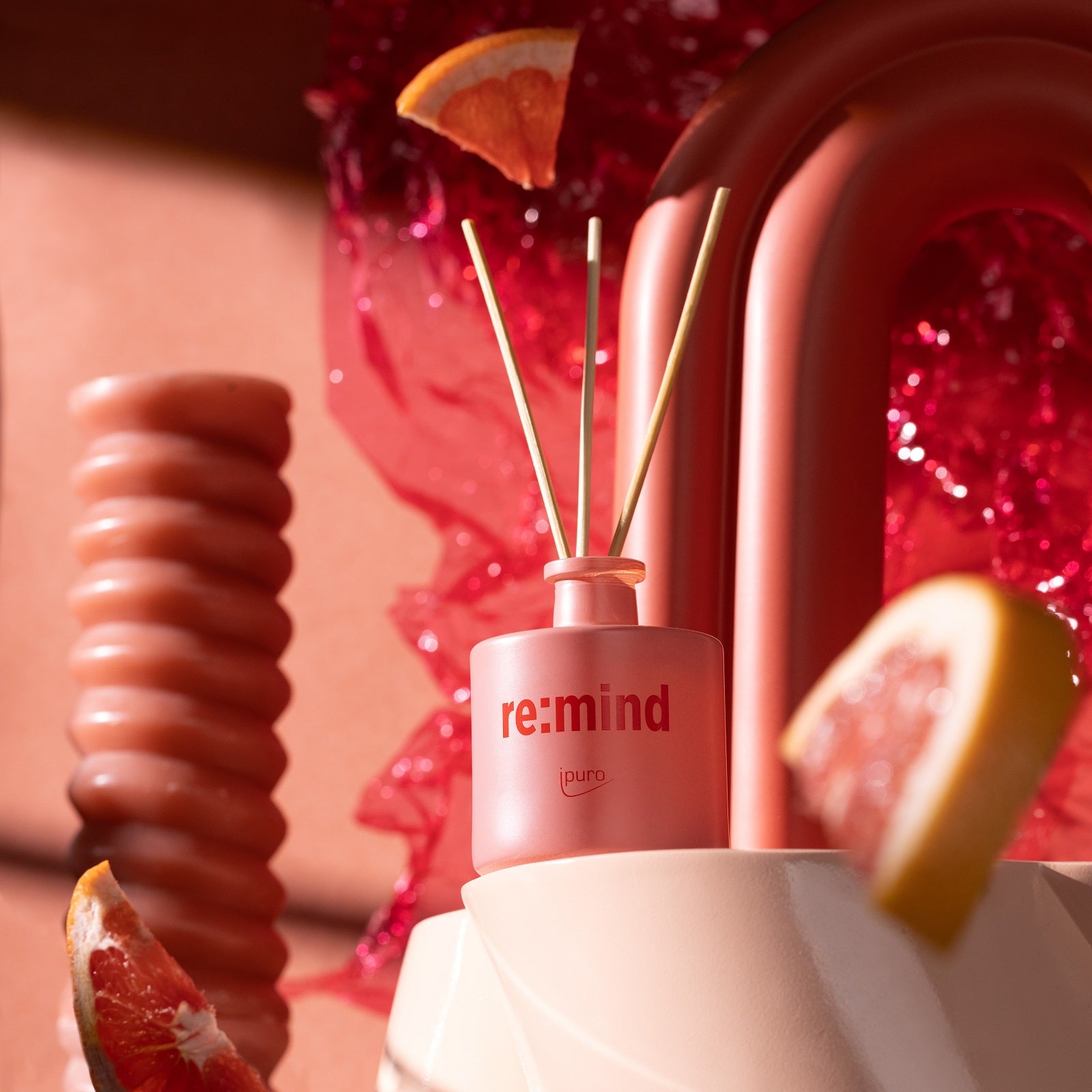 Ein WE ARE: ipuro re:mind Raumduft Diffusor von ipuro steht zwischen abstrakten rosa Skulpturen und Grapefruit-Scheiben, mit einem hellen, modernen Hintergrund und sanftem Sonnenlicht, durchdrungen von erfrischendem Grapefruit-Duft.