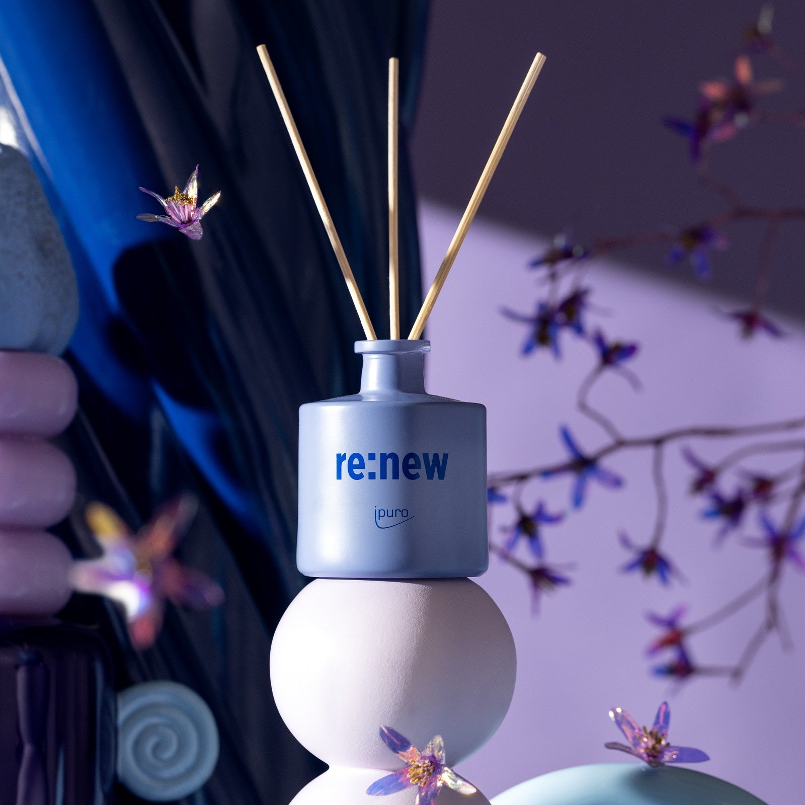 Ein blauer WE ARE: ipuro re:new Raumduft-Flakon mit drei Schilfrohren von ipuro steht auf pastellfarbenen Formen zwischen violetten Blumen und sanfter Beleuchtung und unterstreicht diesen Upcycling-Duft aus geretteten Blumen in einer modernen, künstlerischen Szene.