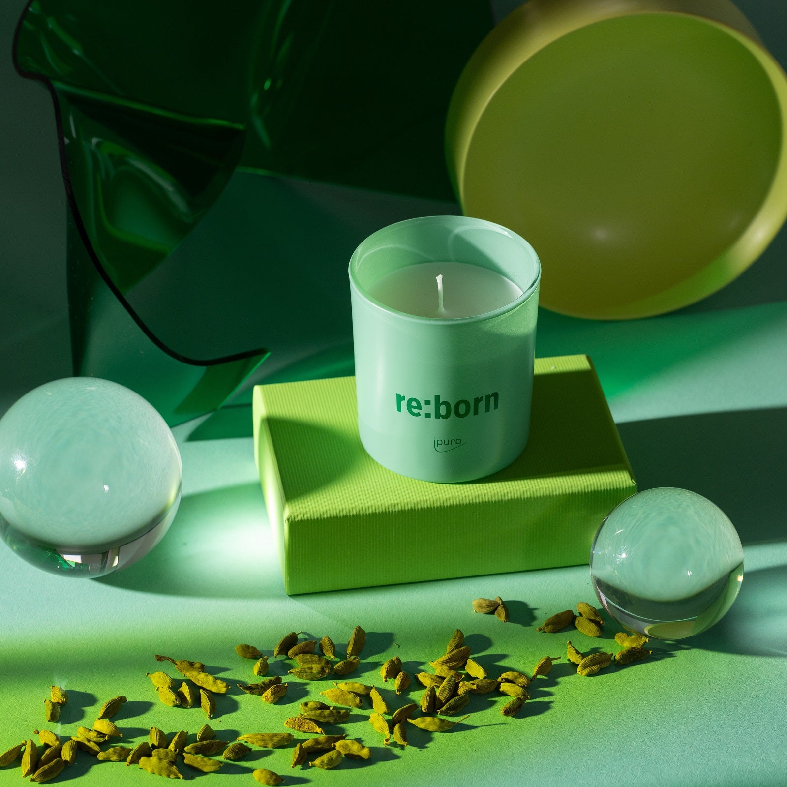 Die WE ARE: ipuro re:born Duftkerze von ipuro, hergestellt aus upgecycelten Gewürzsaaten und tierversuchsfrei, sitzt auf einer grünen Box inmitten von Glaskugeln und Kardamomkugeln - eine dramatische Beleuchtung unterstreicht ihren Raumduft-Charme.