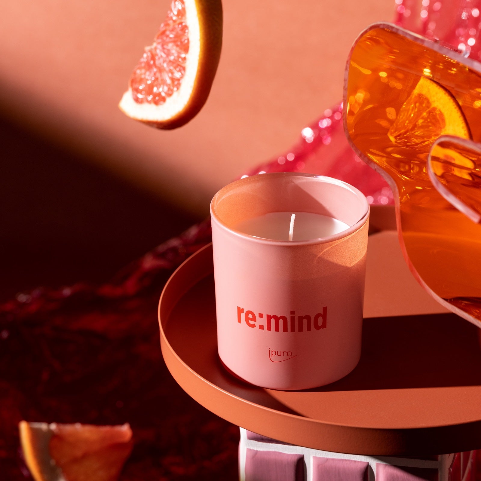 Die WE ARE: ipuro re:mind Duftkerze von ipuro in Pink mit Grapefruitduft wird auf einer runden Terrakottafläche zusammen mit Grapefruitscheiben und einem Glas Orangenflüssigkeit in einer warm beleuchteten Umgebung präsentiert.