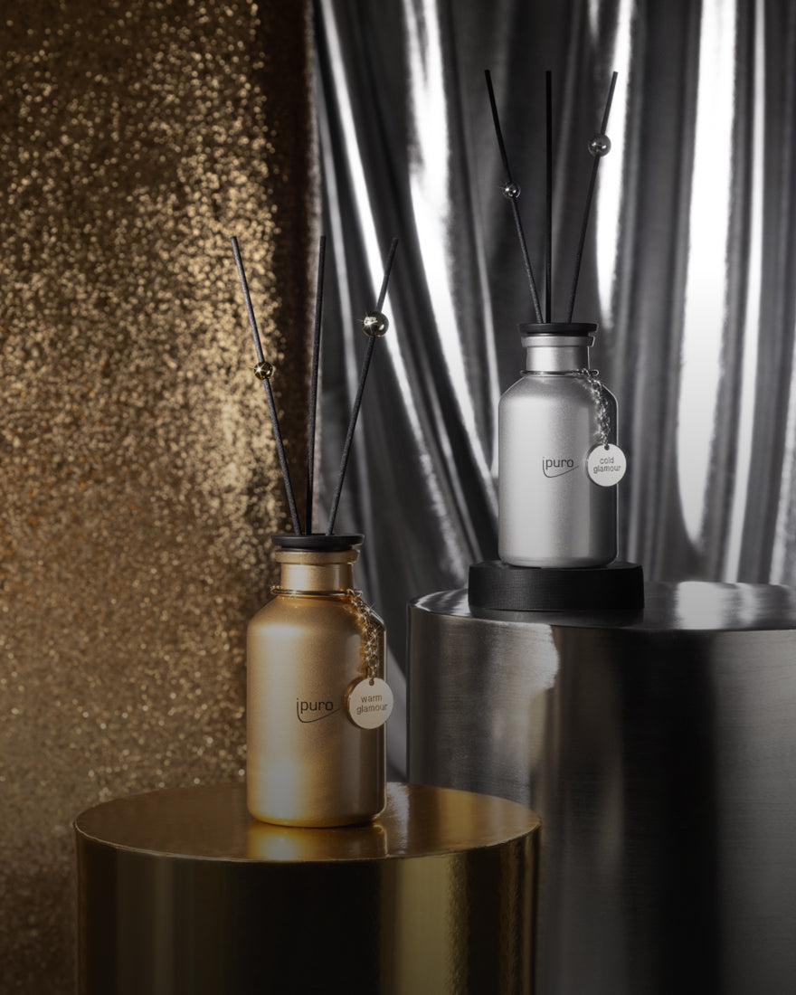 Zwei dekorative Diffusoren in goldenen und silbernen Flaschen mit der Aufschrift "ipuro" werden auf metallischen Sockeln in Gold und Silber ausgestellt, mit einem glänzenden goldenen Paillettenhintergrund und einem silbernen drapierten Stoff im Hintergrund.