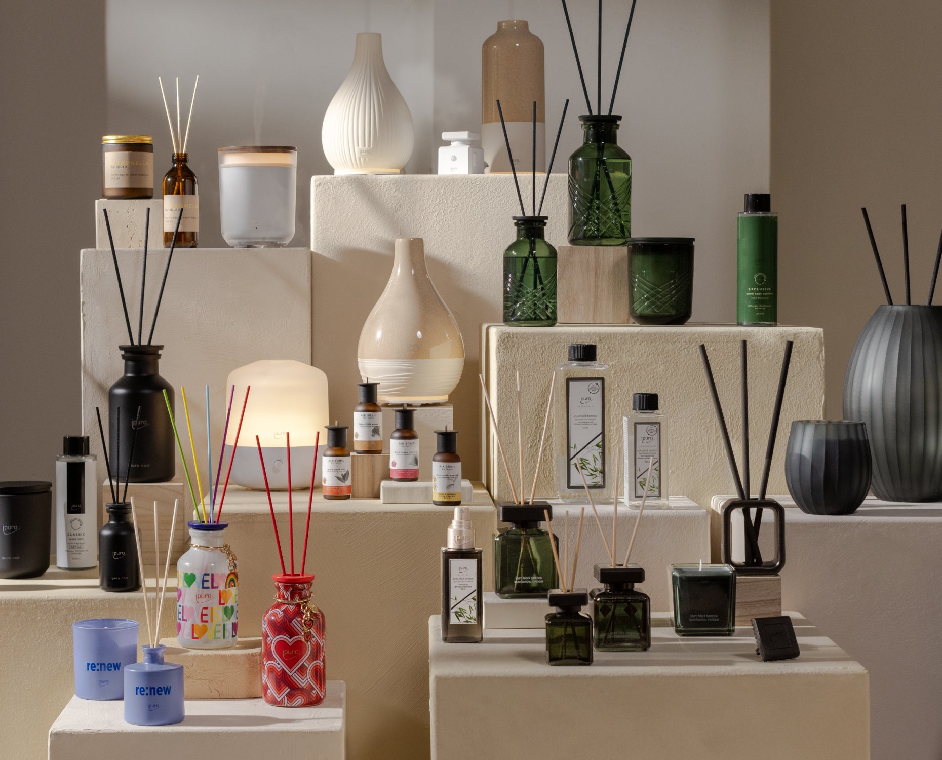 Ein Display mit verschiedenen Duftkerzen, Rohrdiffusoren und dekorativen Vasen auf cremefarbenen Sockeln in einem gut beleuchteten Raum. Die Artikel sind in verschiedenen Formen, Farben und Größen gehalten und bilden ein optisch ansprechendes Arrangement.