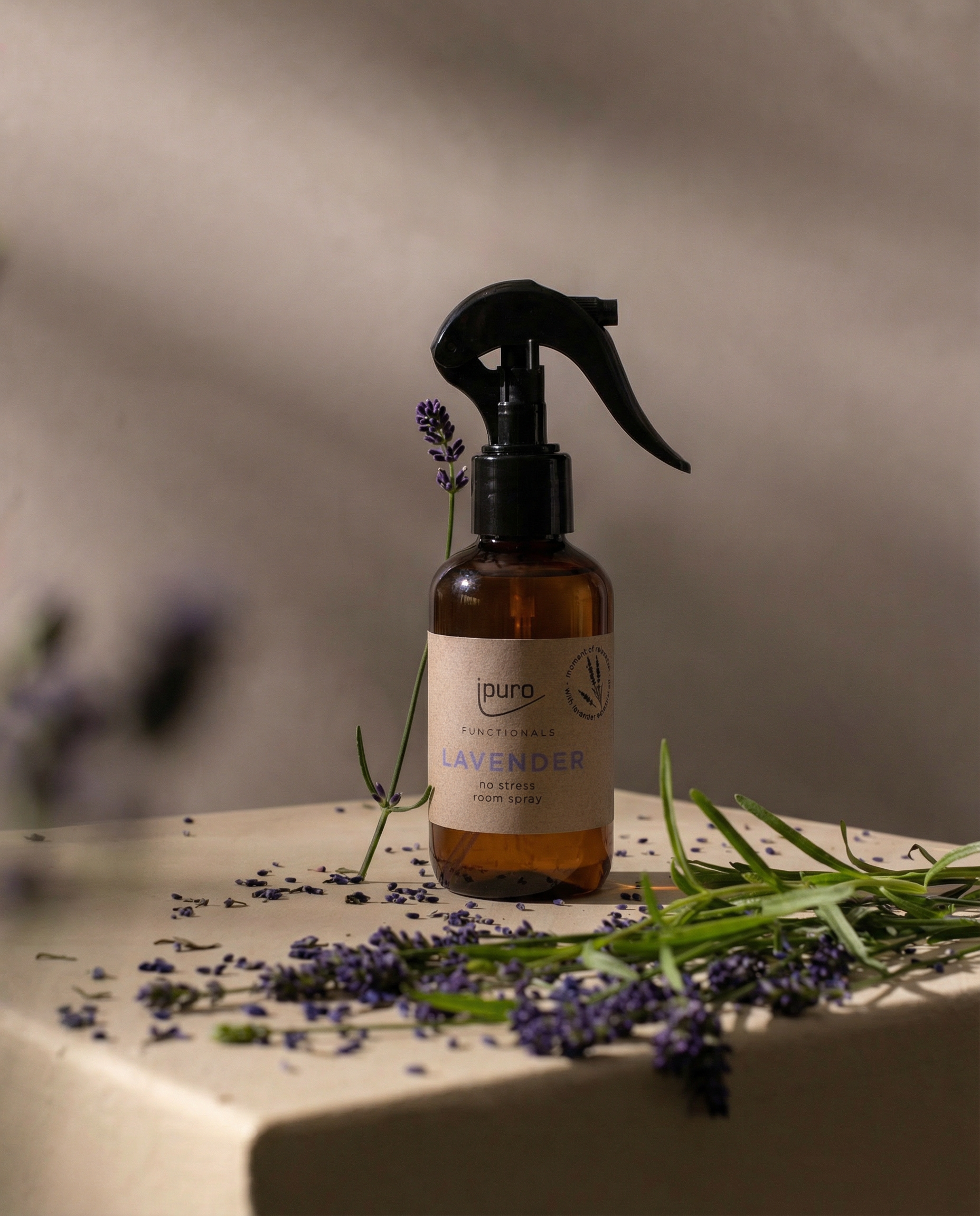 Ein 150ml FUNCTIONALS ipuro lavender Raumspray von ipuro steht auf einer beigen Fläche mit verstreuten Lavendelzweigen und -blüten, beleuchtet von weichem natürlichem Licht vor einem verschwommenen Hintergrund.