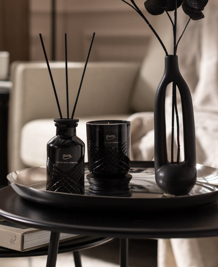 Eine dekorative schwarze Vase mit getrockneten Blumen, eine schwarze Kerze und ein schwarzer Schilfdiffusor stehen auf einem glänzenden runden Tablett auf einem modernen schwarzen Tisch, mit einem beigen Sofa und einem Buch im Hintergrund.