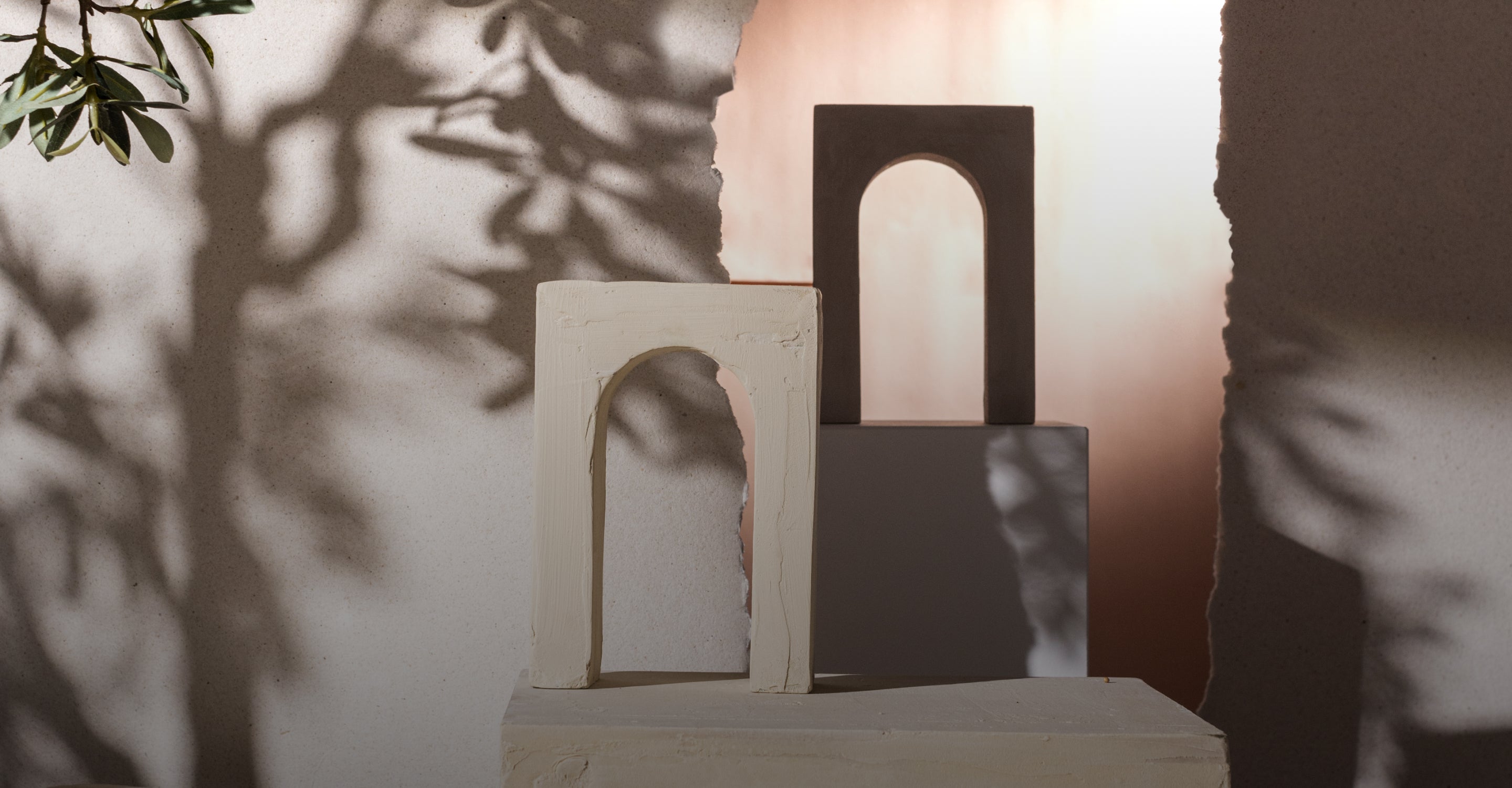 Minimalistische Szene mit zwei Bogenskulpturen, einer cremefarbenen und einer grauen, die auf weißen Podesten stehen. Sanftes Licht wirft blattartige Schatten auf die strukturierte Wand im Hintergrund und schafft eine ruhige, künstlerische Atmosphäre.