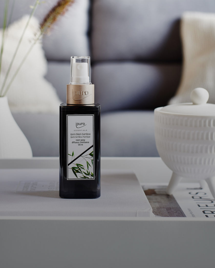 Eine schwarze Flasche ipuro Black Bamboo Diffusorspray mit einer goldenen Kappe steht auf einem weißen Tablett neben einem weißen, strukturierten Behälter in einem modernen, sanft beleuchteten Wohnzimmer.