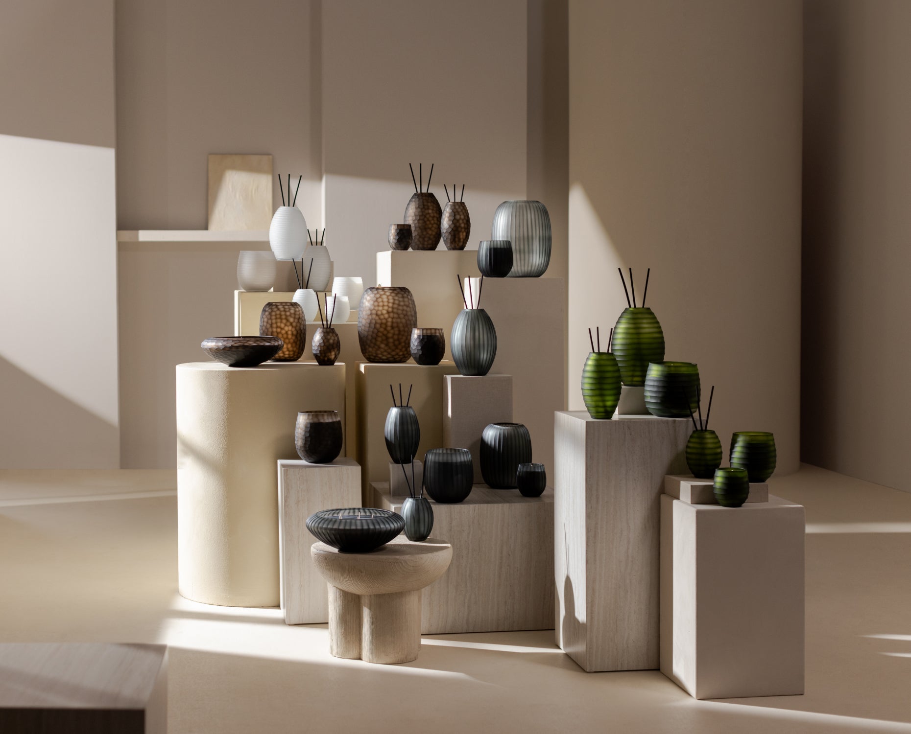 In einem minimalistischen, sonnendurchfluteten Raum mit beigen Wänden sind verschiedene dekorative Vasen und Schälchen in neutralen und grünen Farbtönen auf weißen Sockeln angeordnet.