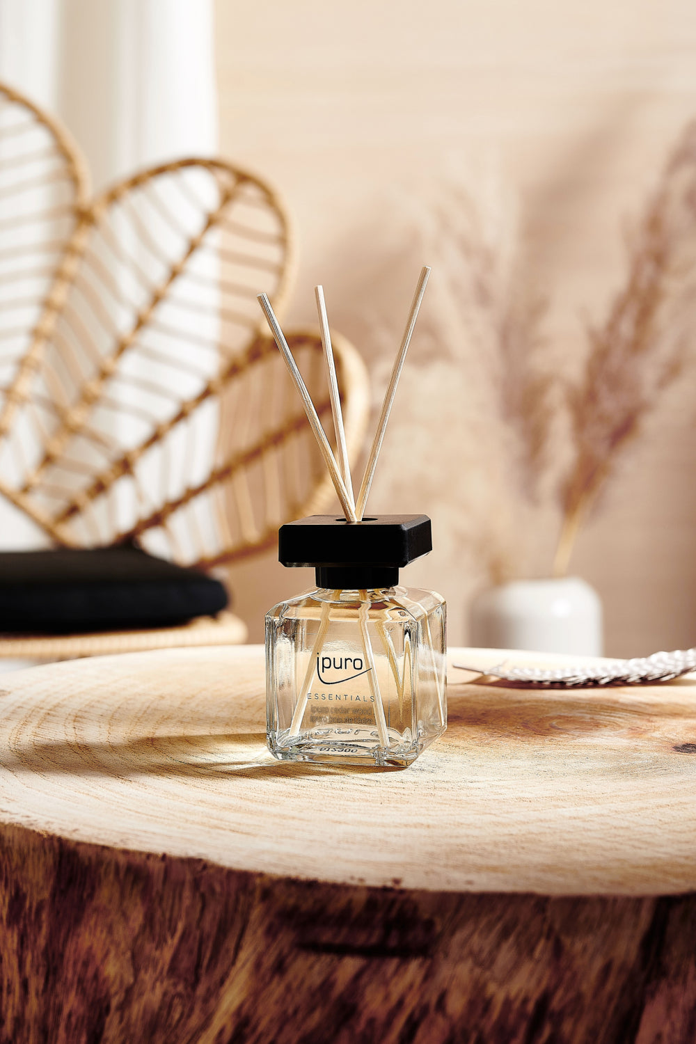 Auf einer hölzernen Unterlage steht ein Glasfläschchen mit der Aufschrift puro ESSENTIAL, in dem sich drei Schilfrohre befinden. Im verschwommenen Hintergrund stehen Rattanmöbel und eine Vase mit getrockneten Gräsern.