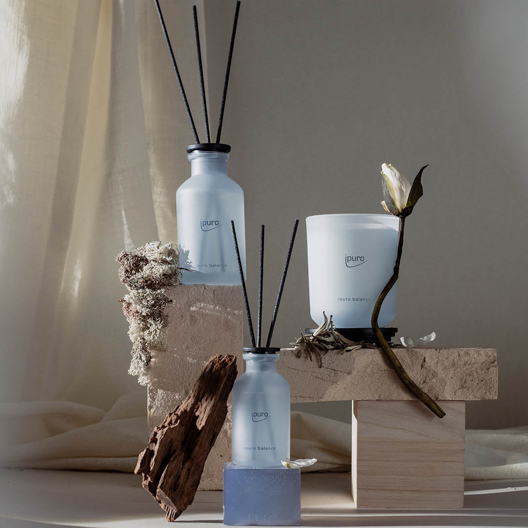 Minimalistische Präsentation des ipuro CLASSIC balance Raumduftes von ipuro, begleitet von einer Kerze und getrockneten Blumen auf Holzblöcken, vor einem neutralen Stoffhintergrund mit subtilen weißen Tee- und maritimen Noten.