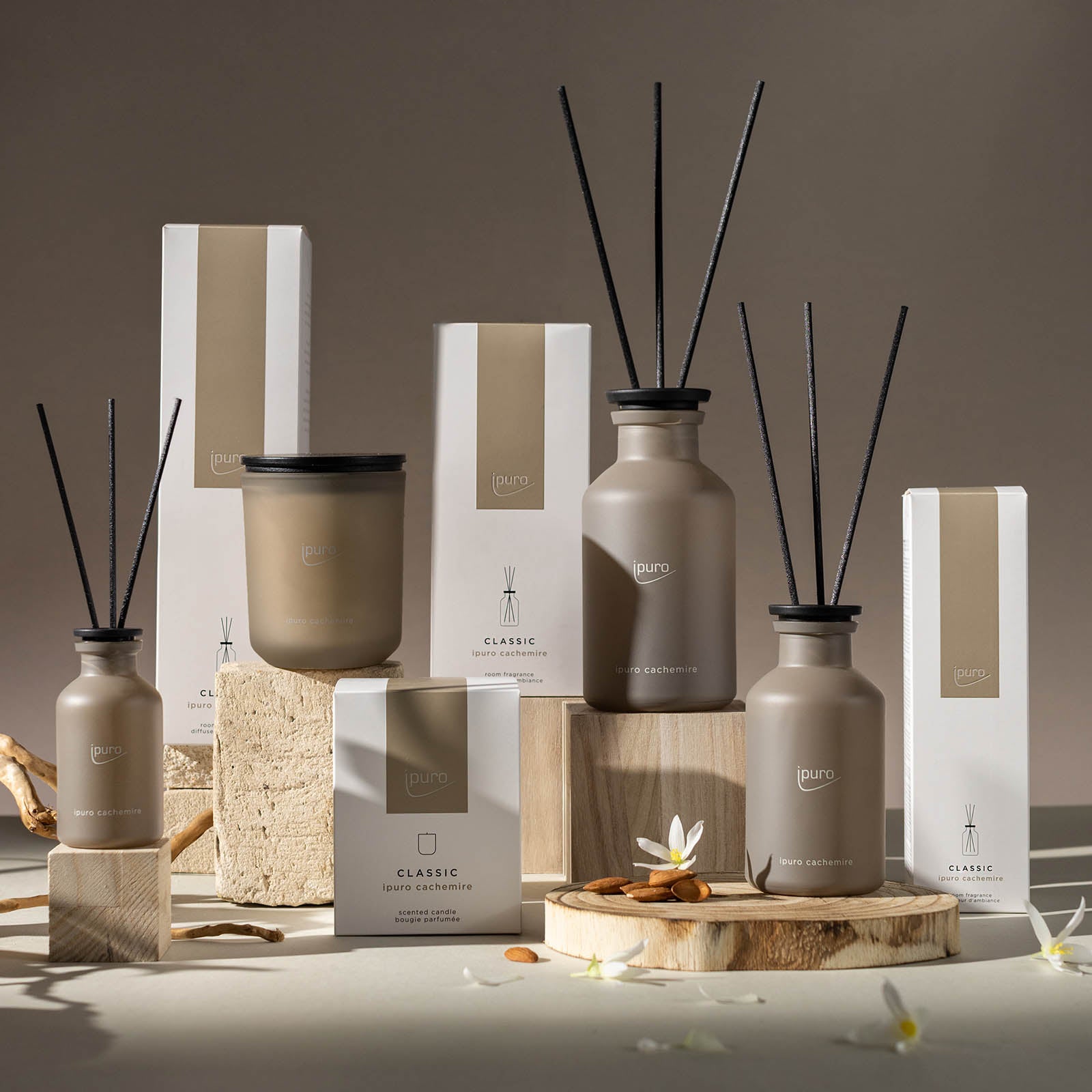 Der CLASSIC ipuro cachemire Raumduft von ipuro, bestehend aus schwarzen Schilfdiffusoren und einer Kerze in neutraler beiger Verpackung, wird elegant auf Holz- und Korkblöcken mit Rosenblättern präsentiert.