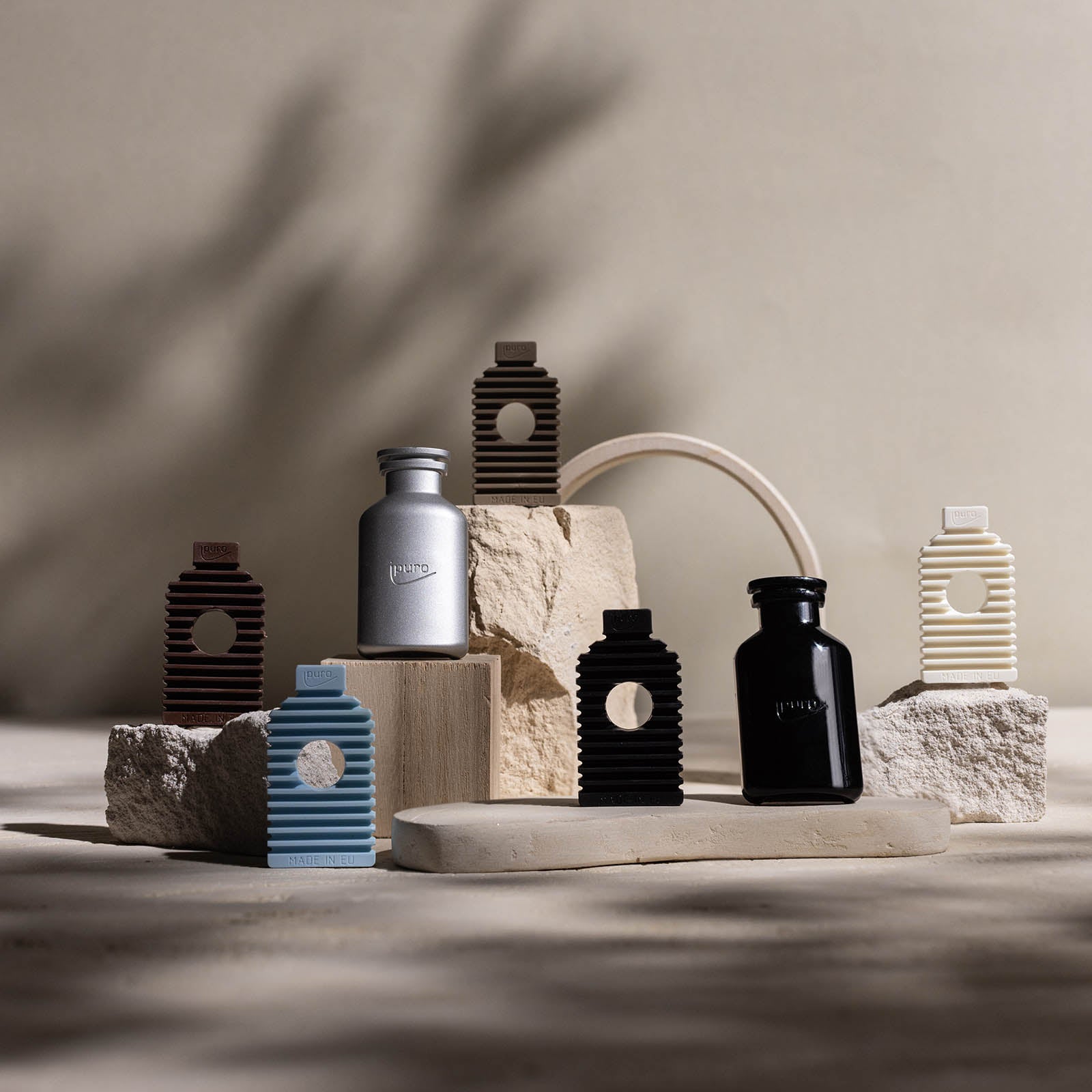 Mehrere kleine, moderne Flaschen und Behälter aus dem ipuro CLASSIC ipuro noir Autoduft Nachfüllpack sind kunstvoll auf strukturierten Steinblöcken angeordnet. Noten von Rose und Sandelholz aus diesem ipuro-Produkt verleihen der minimalistischen Szene Eleganz.