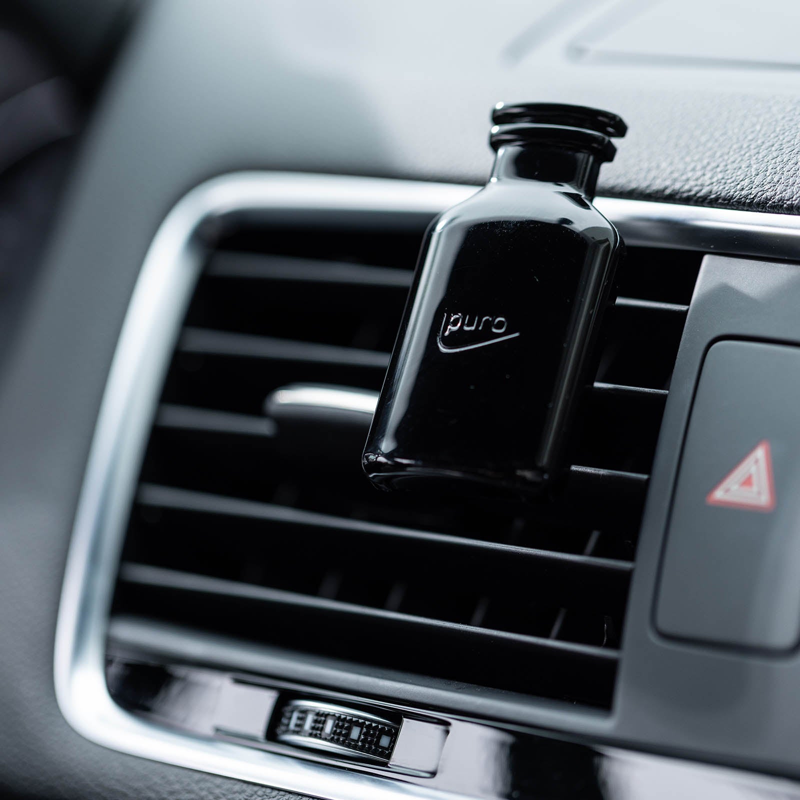 Ein kleiner schwarzer ipuro CLASSIC cuir Autoduft Nachfüllpack ist an der Lüftungsöffnung eines Autos befestigt, wobei die Details des Armaturenbretts und der Lüftungsöffnung im Hintergrund deutlich sichtbar sind.