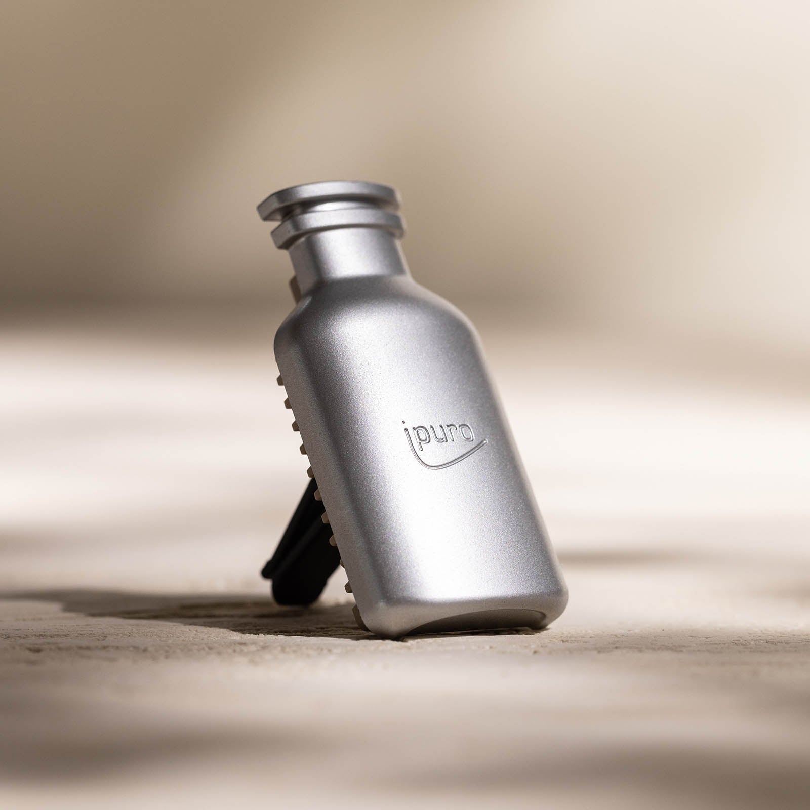 Eine kleine, matt-silberne CLASSIC ipuro blanc Autoduft Nachfüllpack-Flasche von ipuro steht aufrecht auf einer hellen, strukturierten Oberfläche mit weichen Schatten und einem neutralen Hintergrund, der an erfrischende Minze und beruhigende Sandelholznoten erinnert.