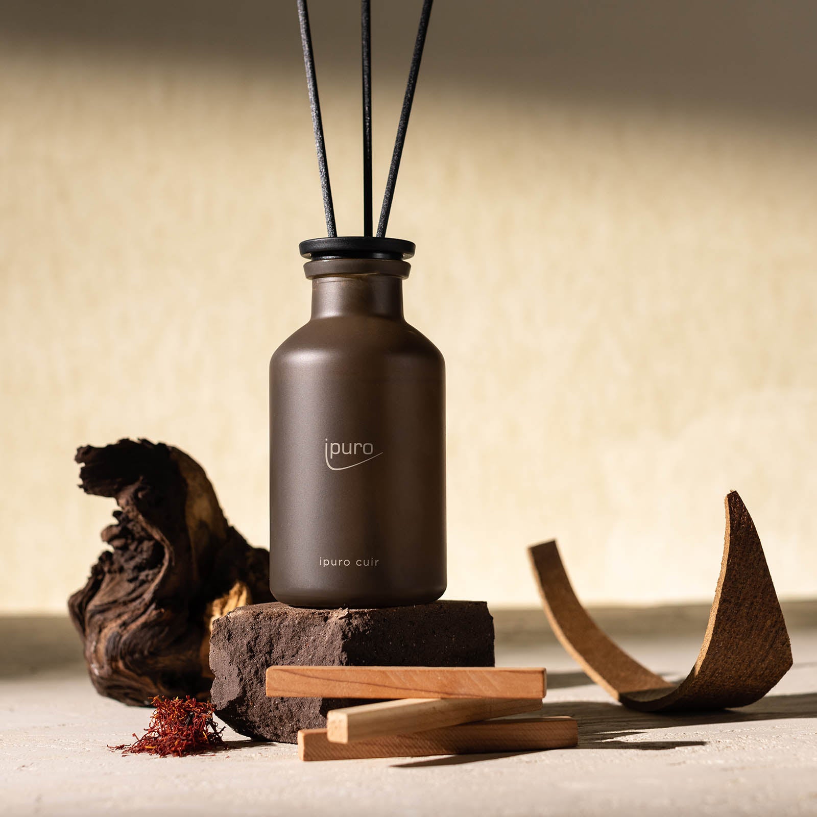 Der CLASSIC ipuro cuir Raumduft von ipuro, ein brauner Glasdiffusor mit schwarzen Stäbchen, ruht auf einer Steinoberfläche mit Holz, Rinde und Safran vor einem beigen Hintergrund. Sein Etikett suggeriert einen eleganten Lederduft mit subtilen Zedernholznoten.