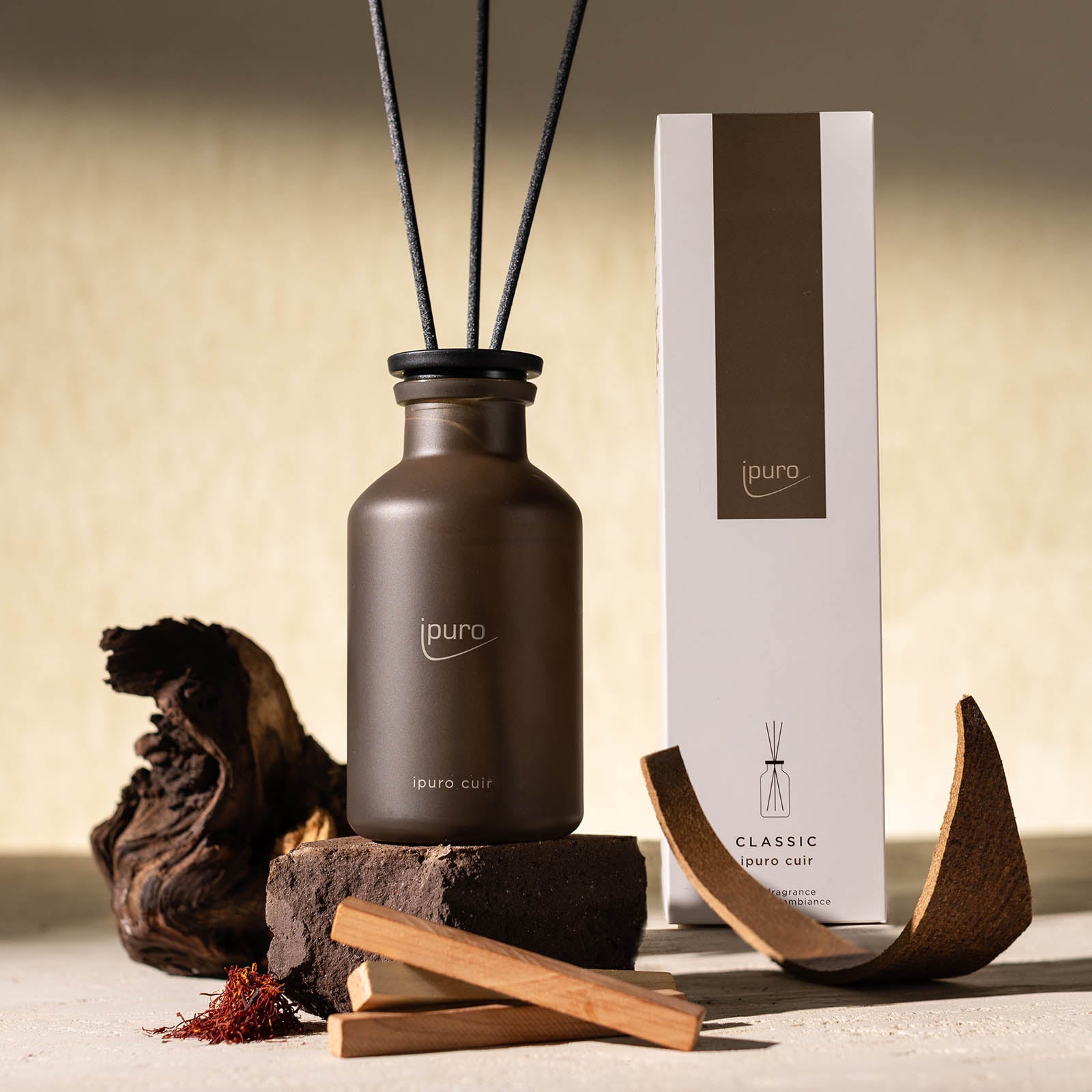 Ein CLASSIC ipuro cuir Raumduft Diffusor von ipuro mit schwarzen Stäbchen wird neben seiner weißen und braunen Verpackung gezeigt, umgeben von Holzstücken, Rinde, Zedernholz und Safranfäden auf einer beigen Oberfläche.