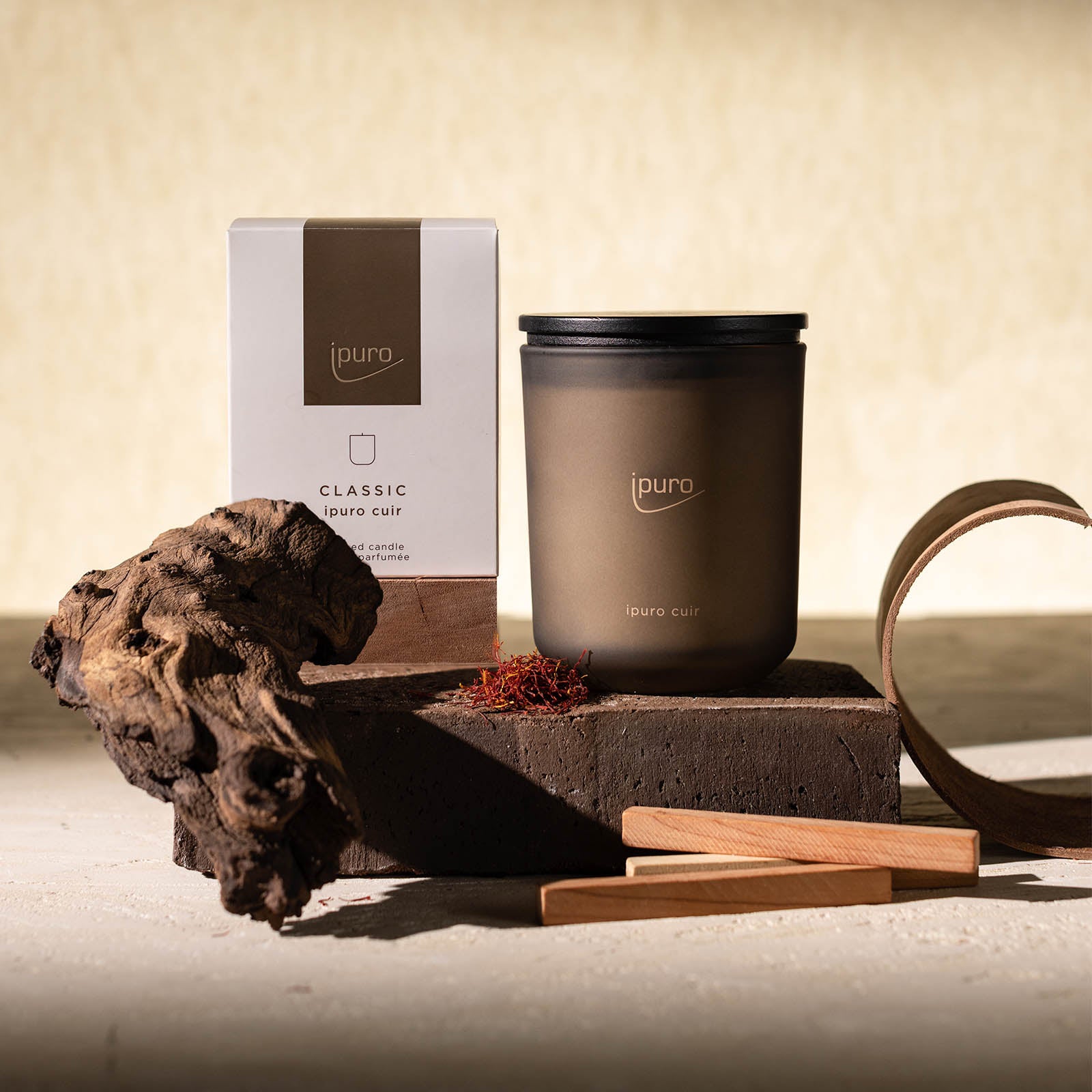 Die CLASSIC ipuro cuir Duftkerze von ipuro, eine Kerze aus braunem Milchglas, steht neben ihrer Dose auf einem Stein mit dekorativem Holz und Stäbchen. Ihre warmen Leder- und subtilen Vanille-Noten schaffen ein raffiniertes, minimalistisches Ambiente.