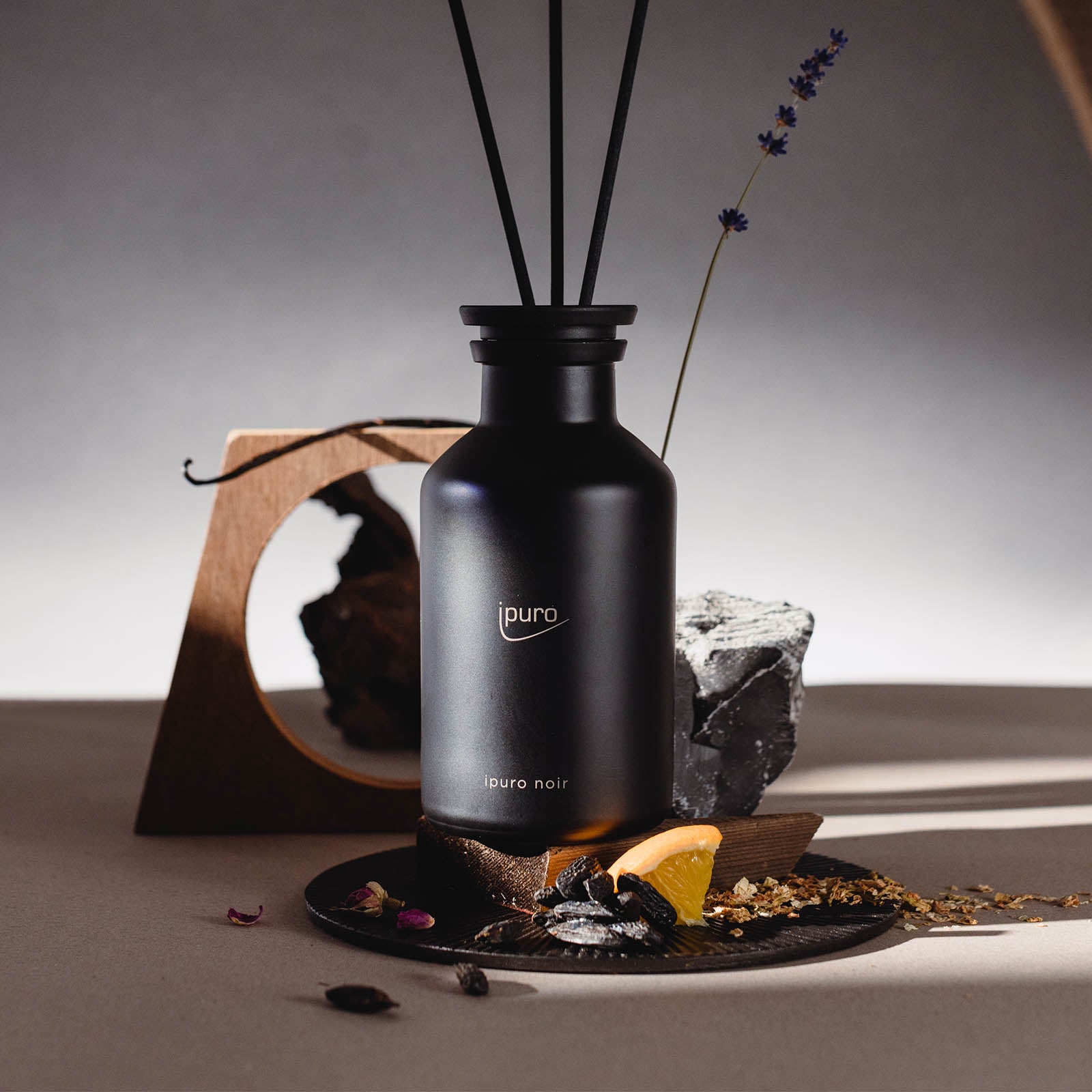 Das CLASSIC ipuro noir Raumduft von ipuro mit schwarzem Stäbchen liegt auf einem Tisch zwischen getrockneten Blumen, Sandelholz, einem Felsen, einer Holzskulptur und einer Orangenscheibe, beleuchtet von einer sanften dramatischen Beleuchtung.