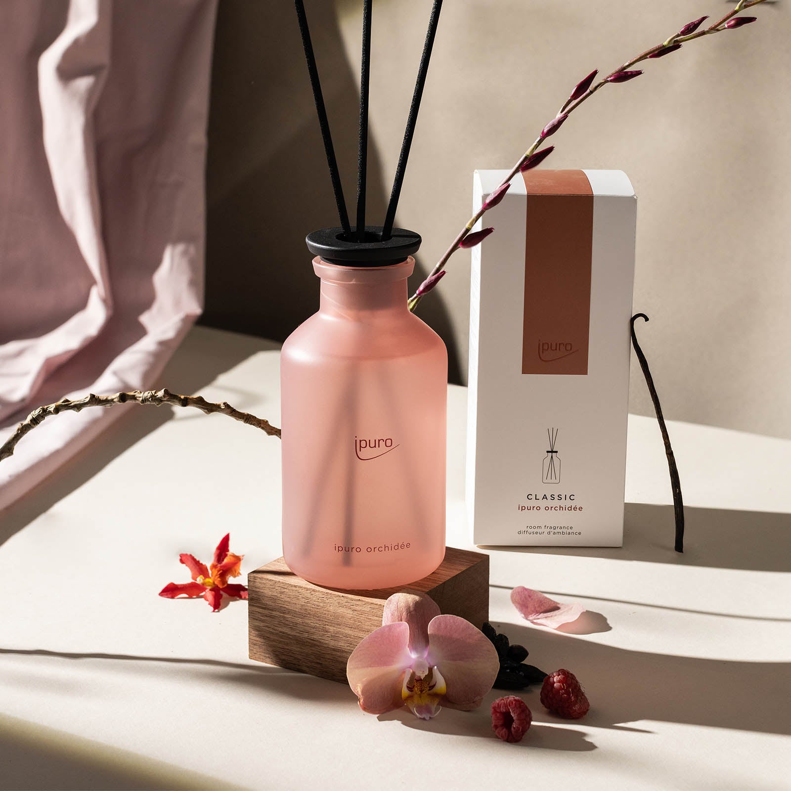 Ein rosafarbener, mattierter Glasflakon von ipuro CLASSIC orchidée Raumduft mit schwarzem Schilfrohr steht auf einem Holzblock inmitten von Blumen und getrockneten Pflanzen, dahinter die passende Schachtel, die an zarte Pfirsichblüten erinnert.
