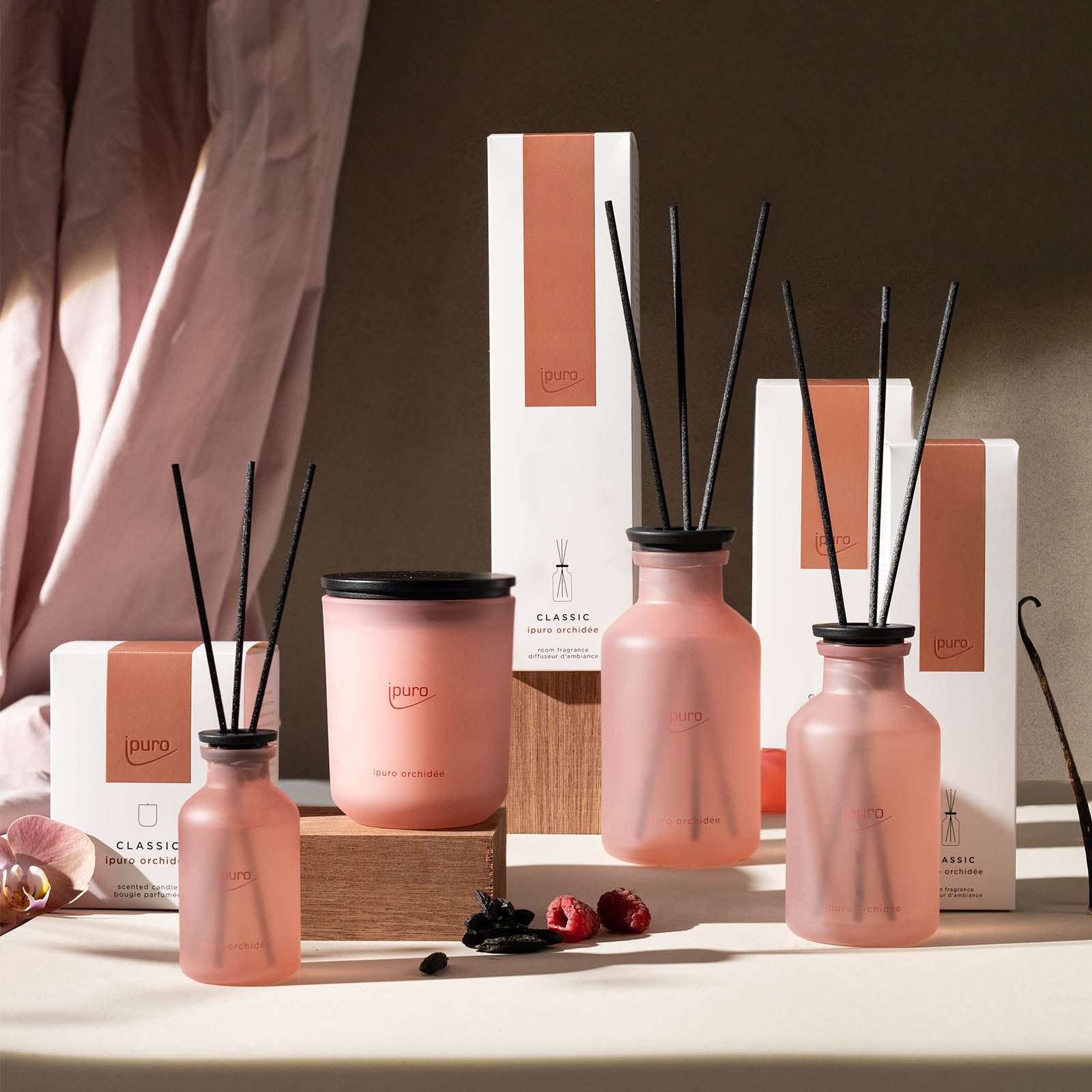 Die CLASSIC ipuro orchidée Duftkerze von ipuro wird mit passenden rosa Milchglasdiffusoren und passender Verpackung auf einer hellen Fläche mit neutralem Stoffhintergrund, Trockenblumen und Holzakzenten präsentiert.