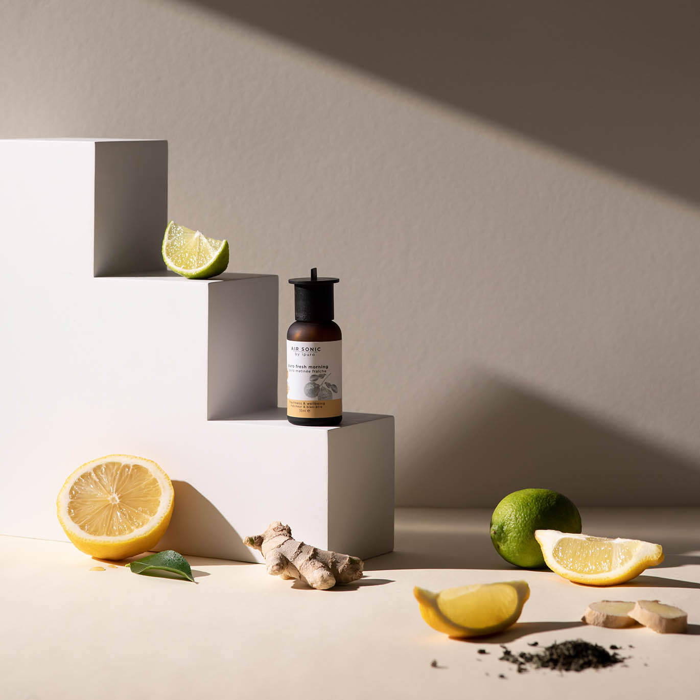Das AIR SONIC ipuro fresh morning Duftöl von ipuro präsentiert sich auf weißen, geometrischen Blöcken mit Zitrone, Limette, Ingwer, Teeblättern und einem Hauch von Bergamotte Frische, beleuchtet von natürlichem Sonnenlicht und weichen Schatten.