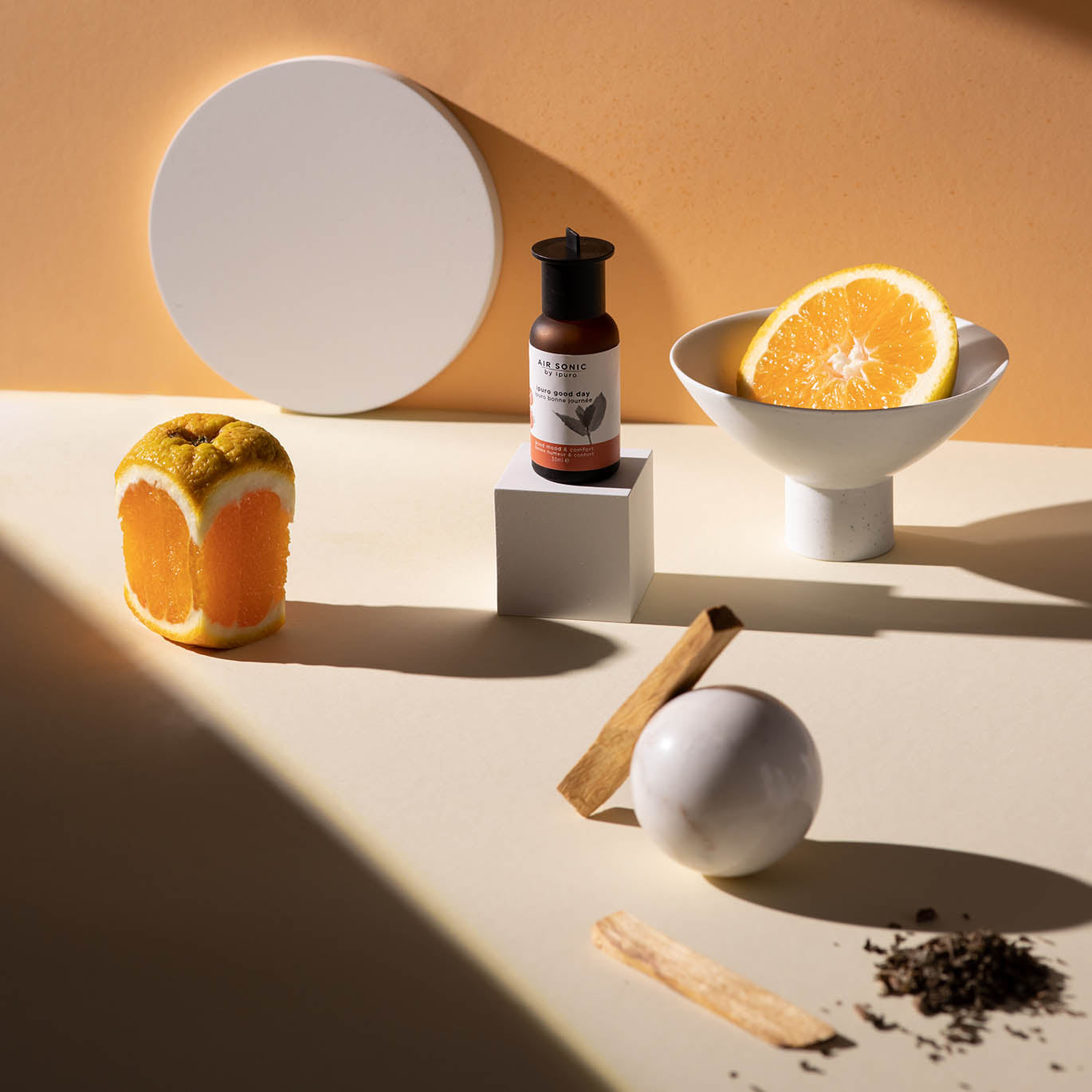Ein Flakon AIR SONIC ipuro good day Duftöl von ipuro steht auf einem weißen Sockel, umgeben von Orangenscheiben, Teeblättern, Holzstäbchen und einem runden weißen Objekt - ideal für Fans der natürlichen Dufttechnologie.