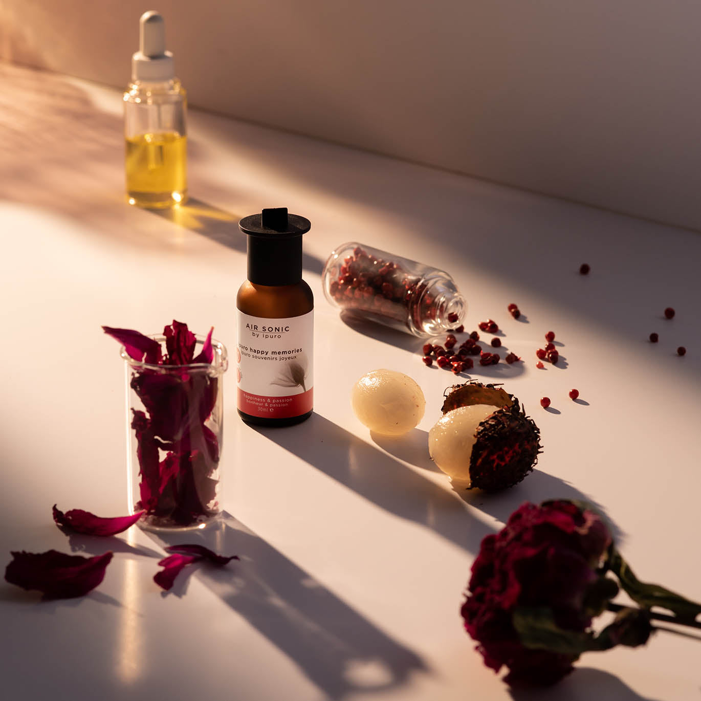 Eine Flasche AIR SONIC ipuro happy memories Duftöl von ipuro, ein Tropfer mit gelber Flüssigkeit, getrocknete Rosenblüten, zwei runde Seifen und eine getrocknete Blume sind auf einer weißen Fläche unter warmer Beleuchtung angeordnet.
