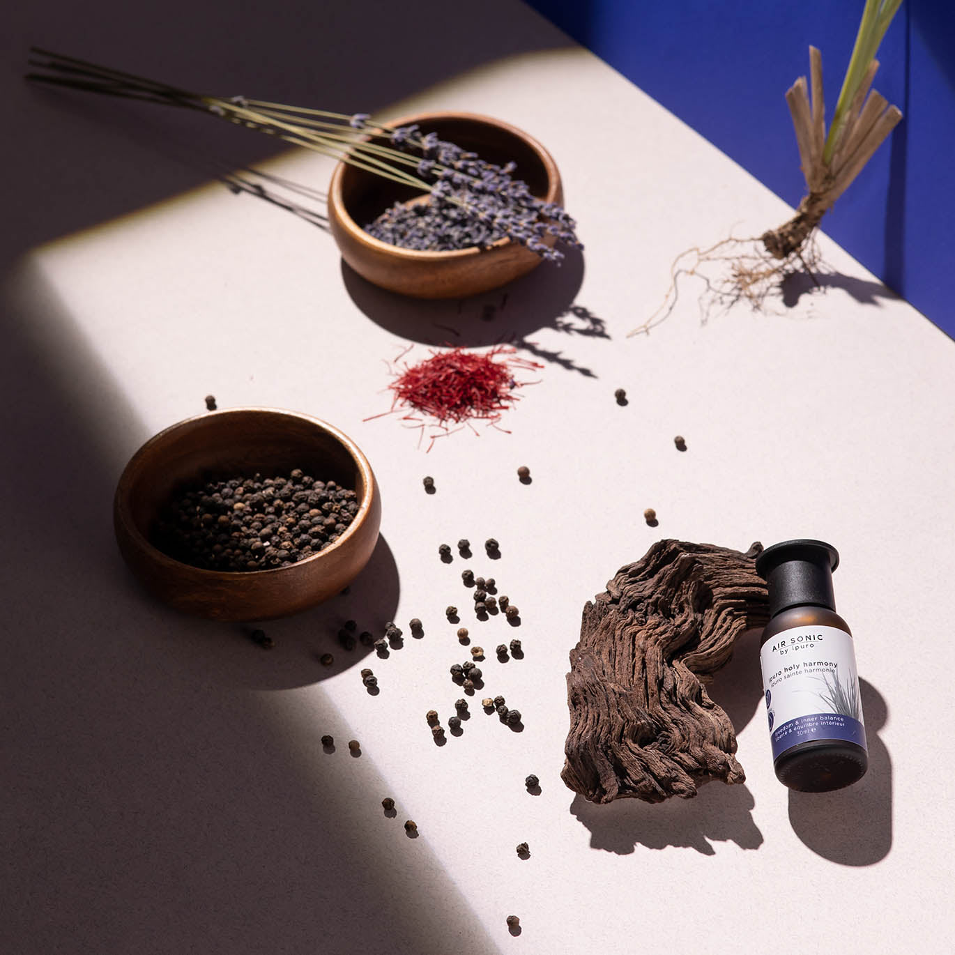 Eine flache Schicht aus pflanzlichen Zutaten - Pfefferkörner, getrockneter Lavendel, rote Safranfäden, Zitronengras, Vetiver aus Haiti, ein strukturiertes Holzstück und ipuro AIR SONIC ipuro holy harmony Duftöl - auf einer hellen Fläche mit Sonnenlicht und blauen Schatten für die innere Balance.
