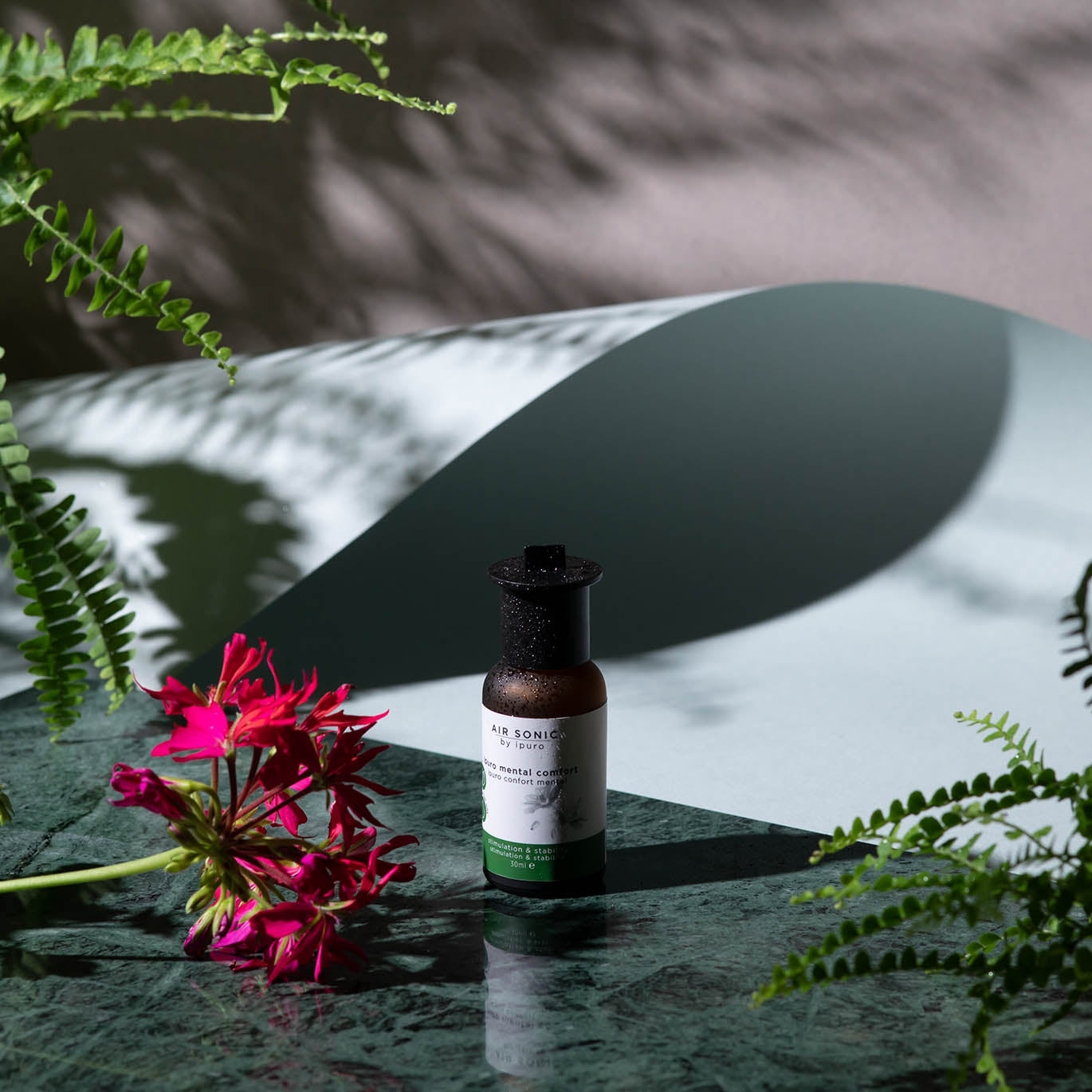 Das AIR SONIC ipuro mental comfort Duftöl von ipuro sitzt auf einer grünen Marmorfläche zwischen Farnen und einer roten Blume, dramatische Licht- und Schattenspiele evozieren mentale Ausgeglichenheit und tiefe Entspannung.