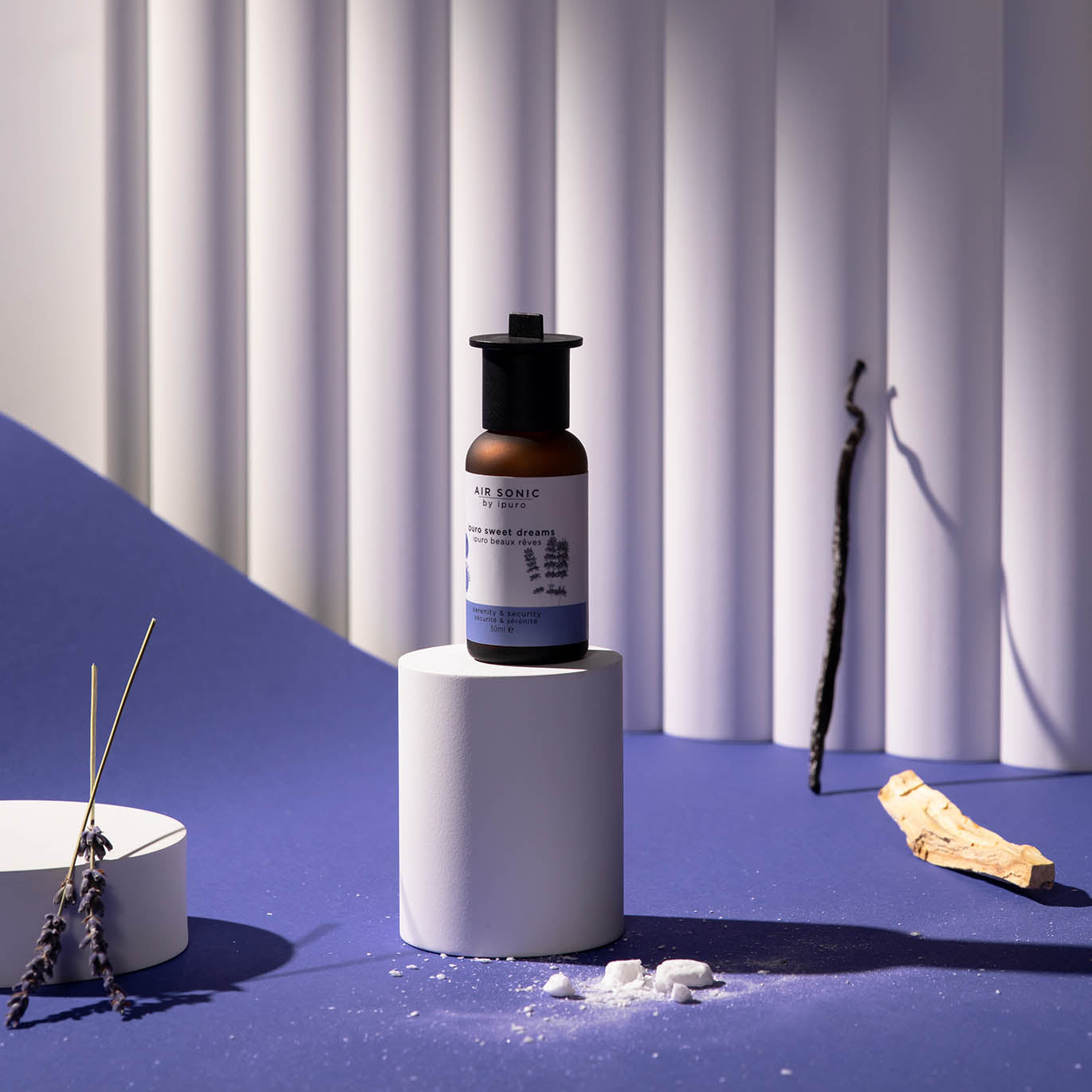 Eine braune AIR SONIC ipuro sweet dreams Duftölflasche mit weißem Etikett steht auf einem weißen Sockel, umgeben von Räucherstäbchen, Lavendelzweigen, weißen Steinen und Holzstücken vor einem blau-weißen abstrakten Hintergrund.