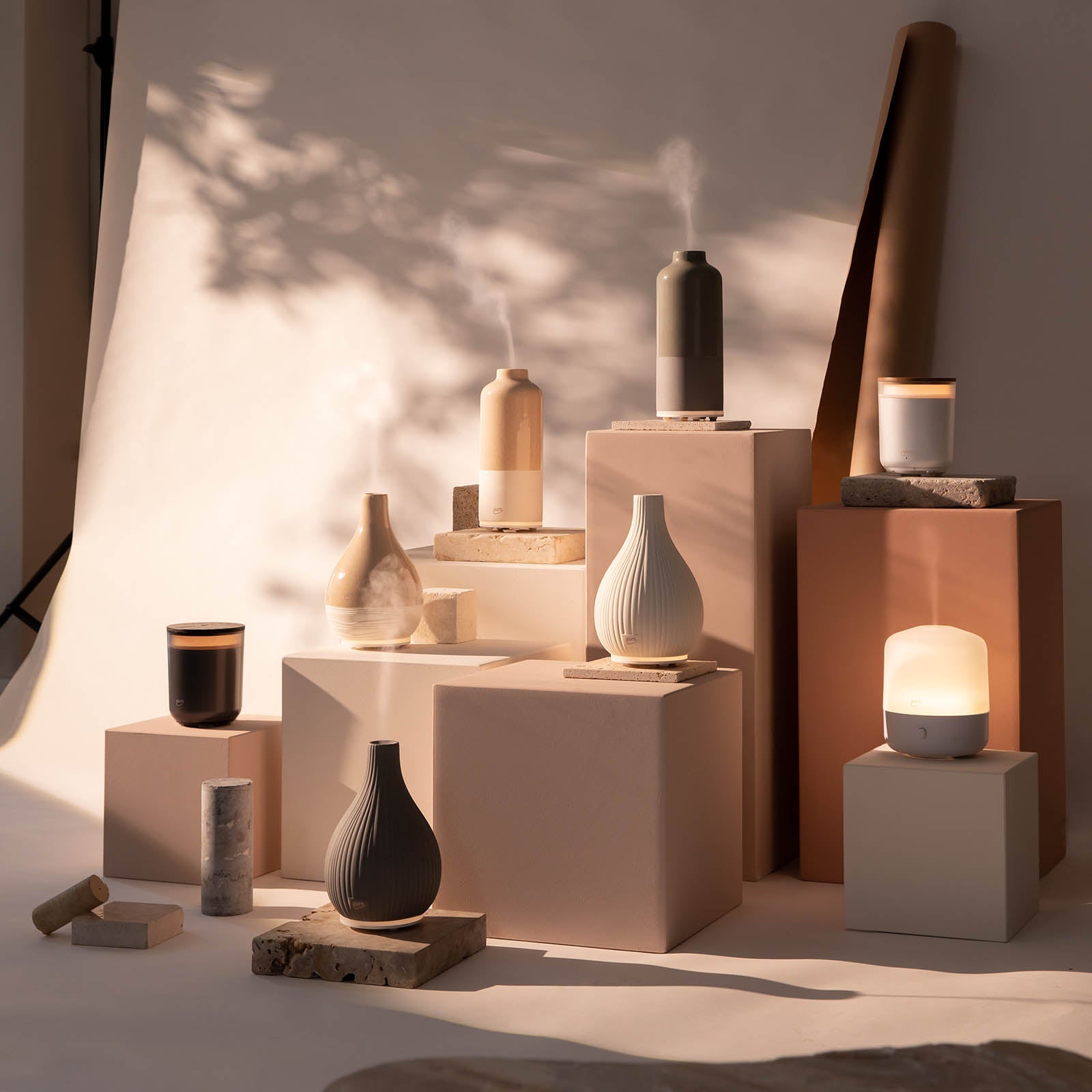 Die ipuro AIR SONIC Aroma-Vase Elektrischer Aroma-Diffusor und Kerzen in neutralen Farbtönen von ipuro stehen auf beigen und weißen Sockeln, die von sanftem Sonnenlicht beleuchtet werden und eine beruhigende Wirkung haben.