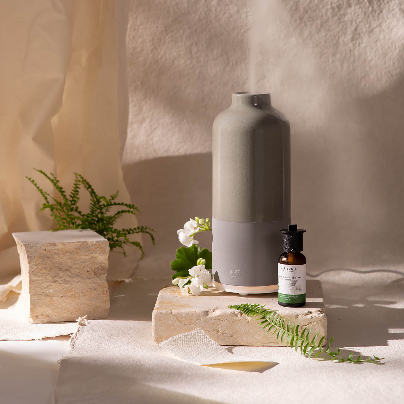 Der AIR SONIC ipuro aroma bottle Elektrischer Aroma-Diffusor von ipuro vernebelt Duftöle als feinen Nebel, steht auf Steinplatten, umgeben von einem braunen Fläschchen, weißen Blumen und Farnen und wird sanft vom Tageslicht beleuchtet.