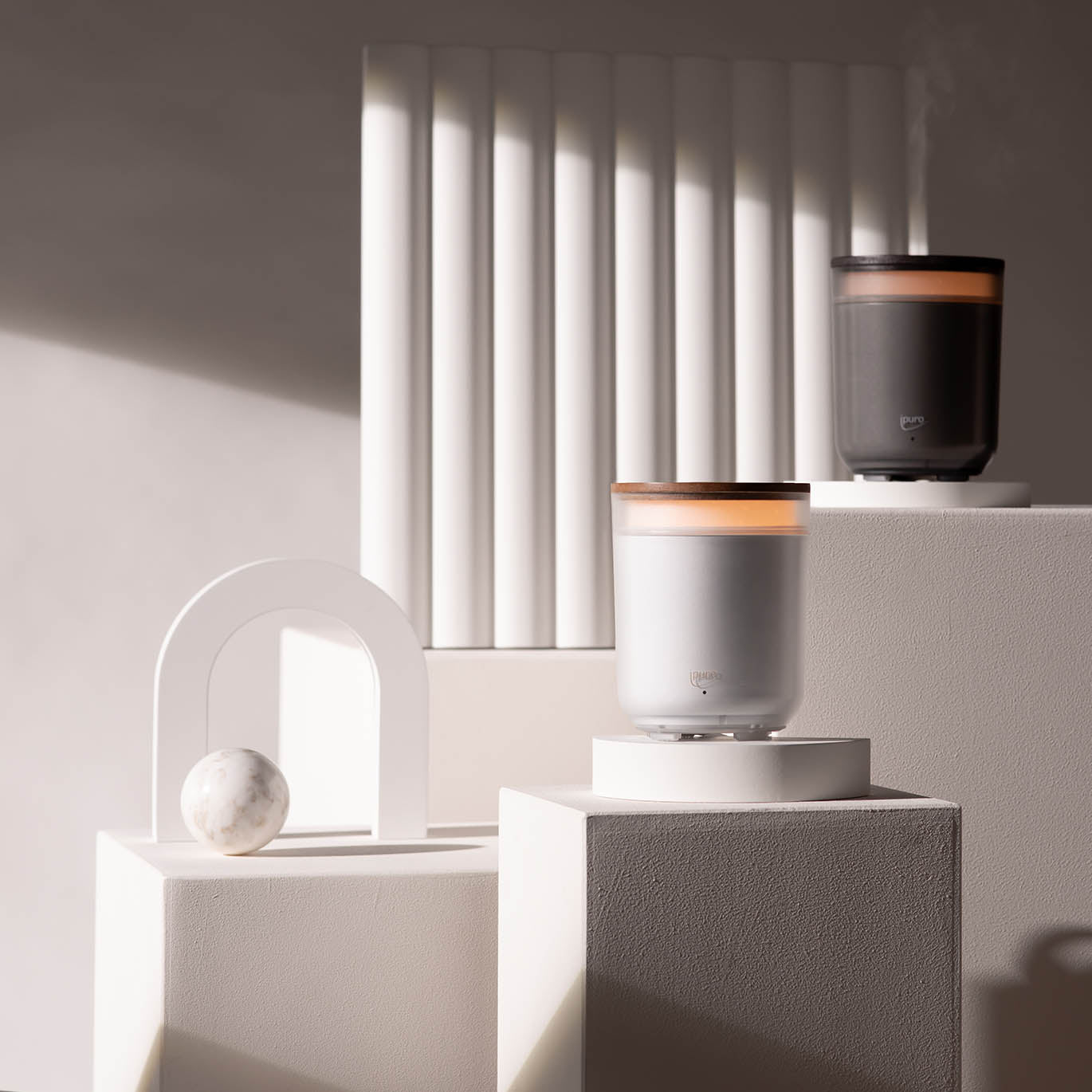 Zwei AIR SONIC ipuro Aromakerzen Elektrischer Aroma-Diffusor von ipuro, ein weißer und ein dunkelgrauer, stehen auf weißen Sockeln mit geometrischem Dekor und einer Marmorkugel. Aus dem dunkelgrauen Diffusor steigt Dampf in einer minimalen, sonnenbeschienenen Umgebung mit vertikalen Rillen auf.