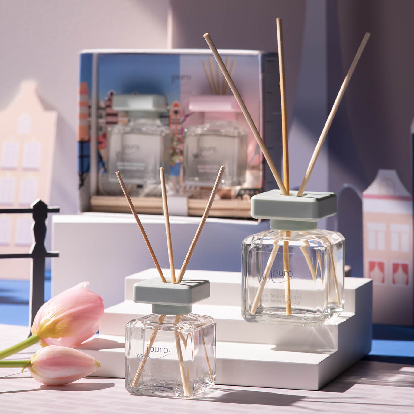Zwei ESSENTIALS ipuro floral amsterdam Diffusoren mit Holzstäben ruhen auf weißen Plattformen neben rosa Tulpen, einer Schachtel und einem illustrierten Stadtbild. Inspiriert vom Amsterdamer Blumenmarkt unterstreicht das sanfte Sonnenlicht die ipuro-Szene.