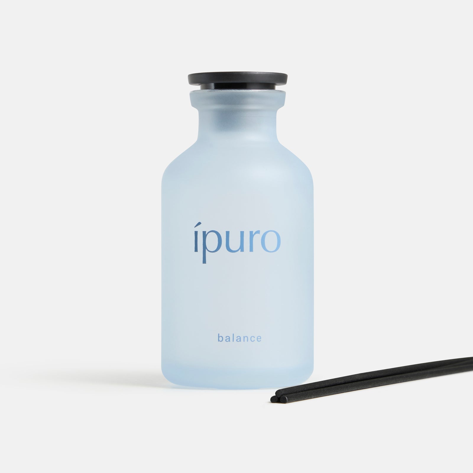 Ein hellblauer Milchglasflakon mit schwarzer Kappe und der Aufschrift ipuro balance Raumduft by ipuro steht neben zwei schwarzen Diffusorstäbchen auf weißem Grund und erinnert an Leichtigkeit und Ruhe.